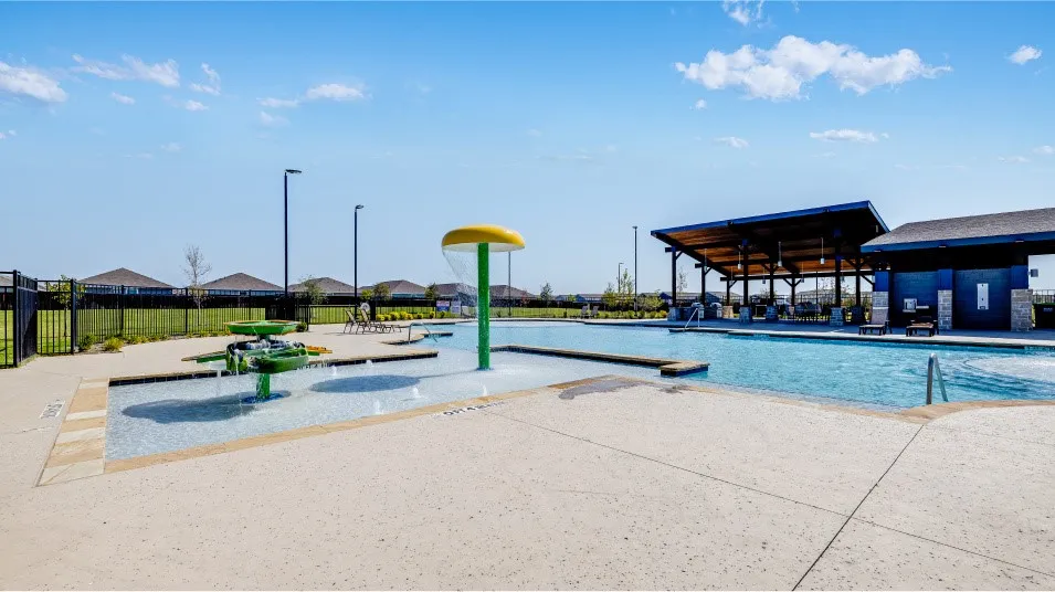 Resort-style aquatic amenity featuring a large swimming pool, splash pad with water features, expansive concrete decking, and an open-air pavilion