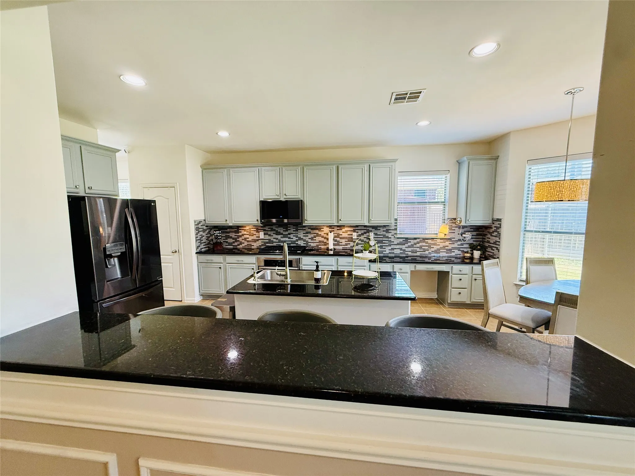 Kitchen featuring an island with a built-in sink, black granite countertops, light gray cabinetry, a mosaic tile backsplash, and recessed lighting