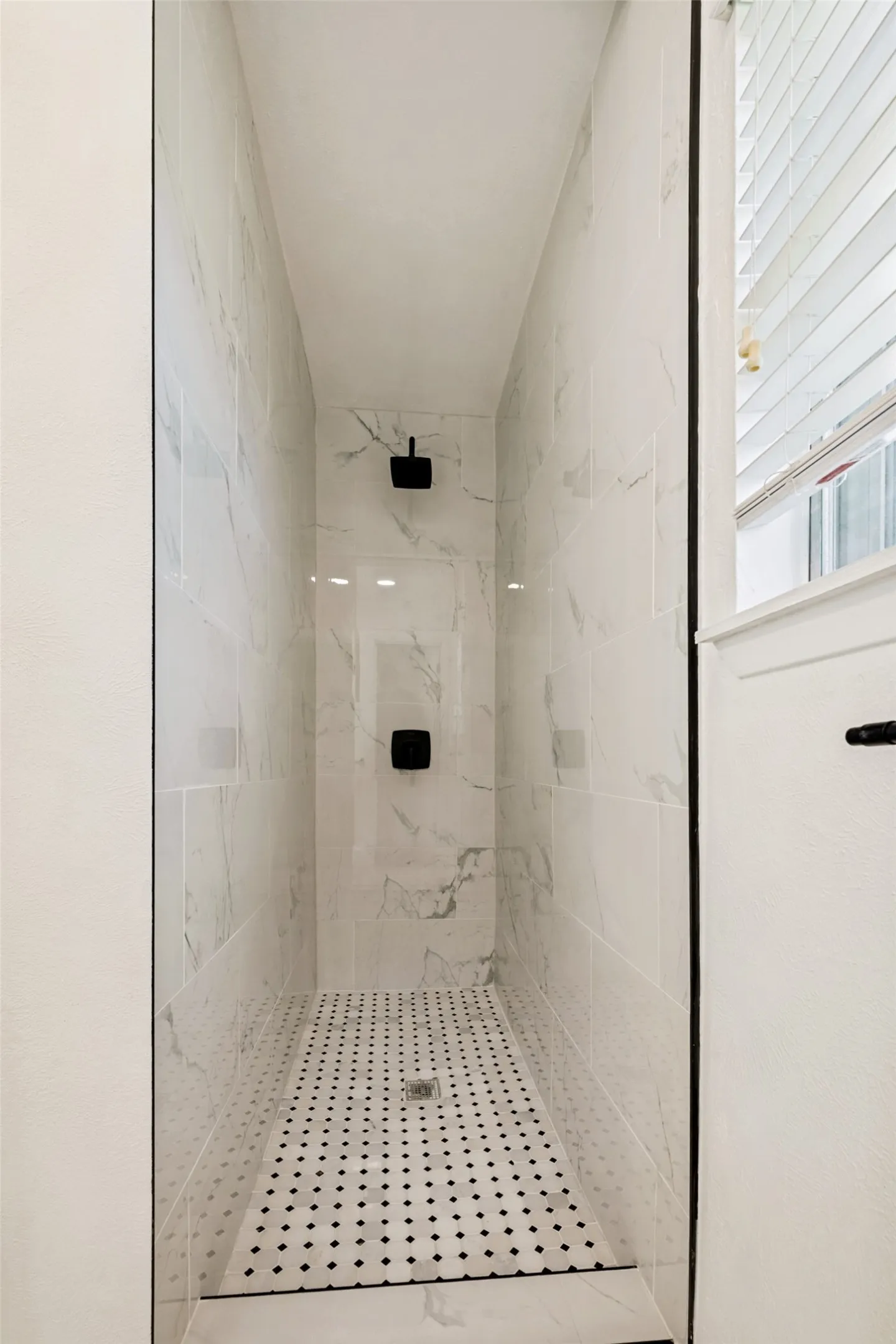 Walk-in shower featuring white marble-look subway tile walls, black and white mosaic tile flooring, and matte black fixtures