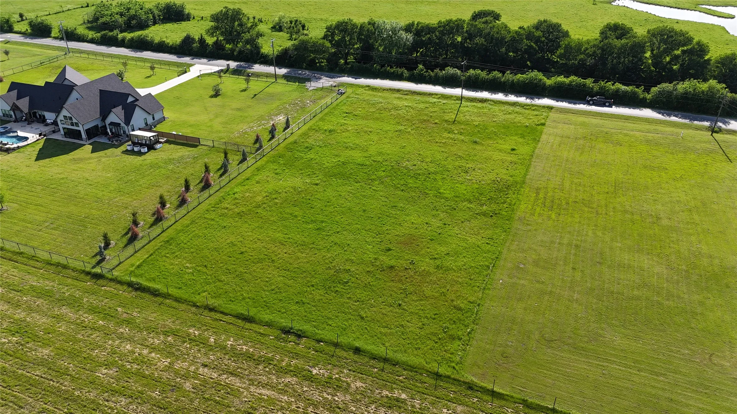 Expansive green property featuring a partially fenced boundary, a paved road border, and distant natural waterways