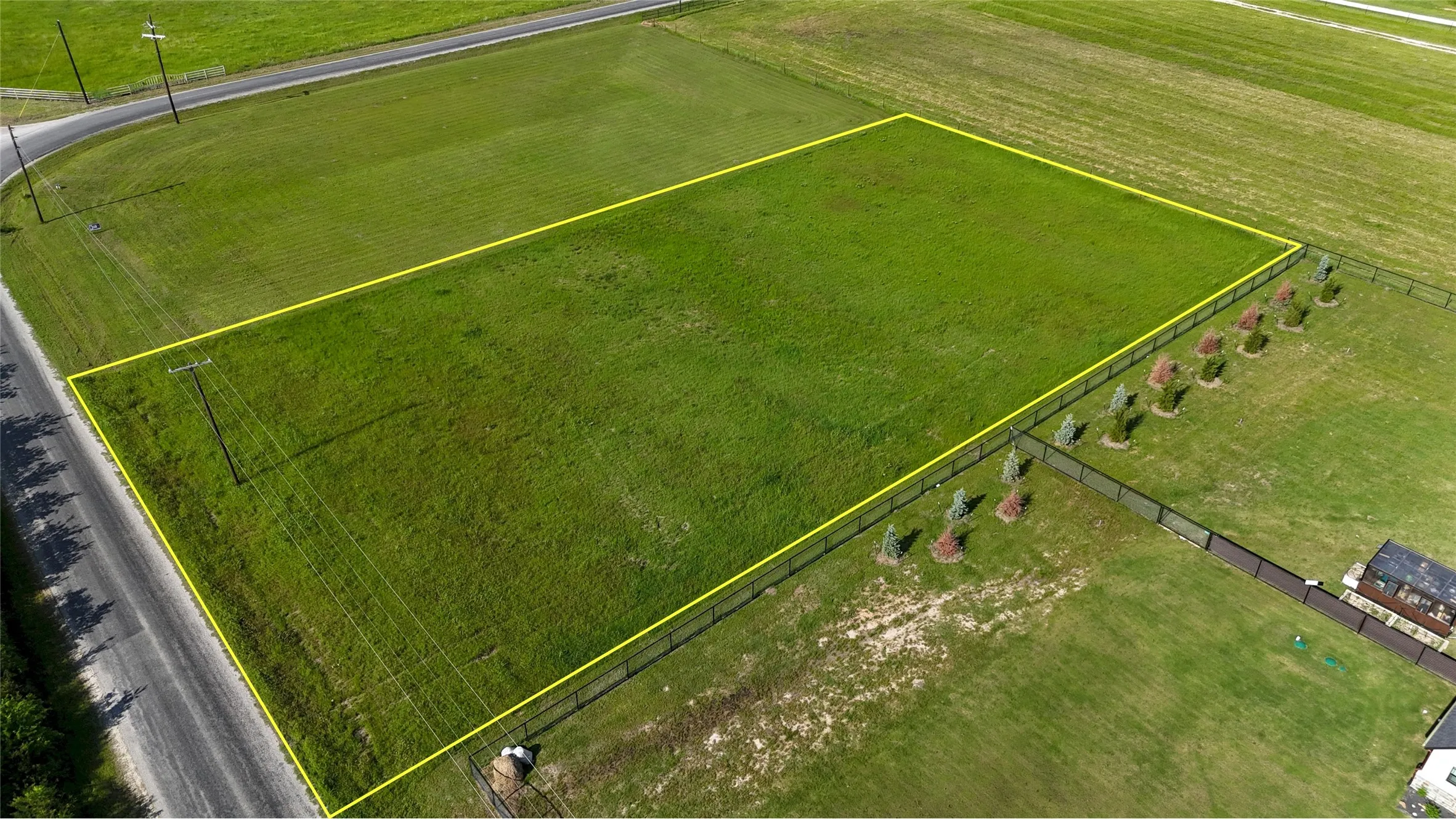 Expansive rectangular lot featuring a manicured grass surface, a black metal perimeter fence, and young trees planted along one boundary