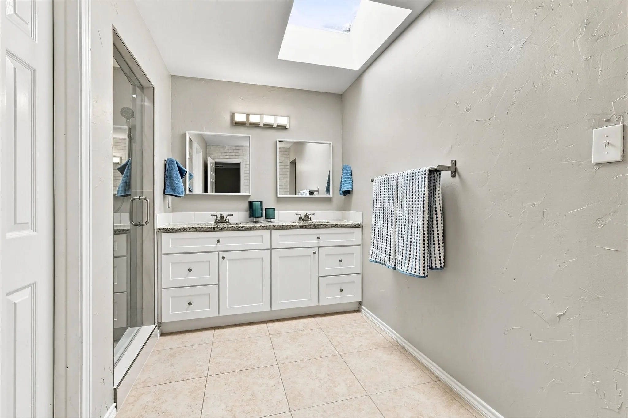 Dual vanity with white cabinetry, a skylight, tile flooring, and a glass-enclosed shower