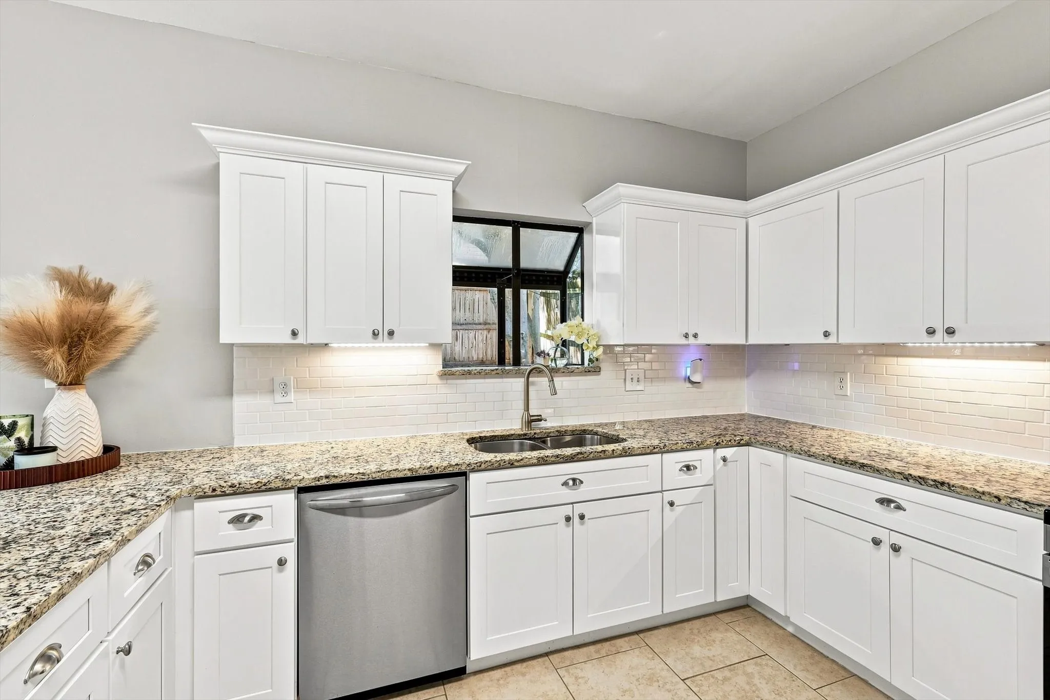 White shaker-style cabinetry with crown molding, granite-finish countertops, subway tile backsplash, stainless steel dishwasher, and a dual basin sink with a gooseneck faucet