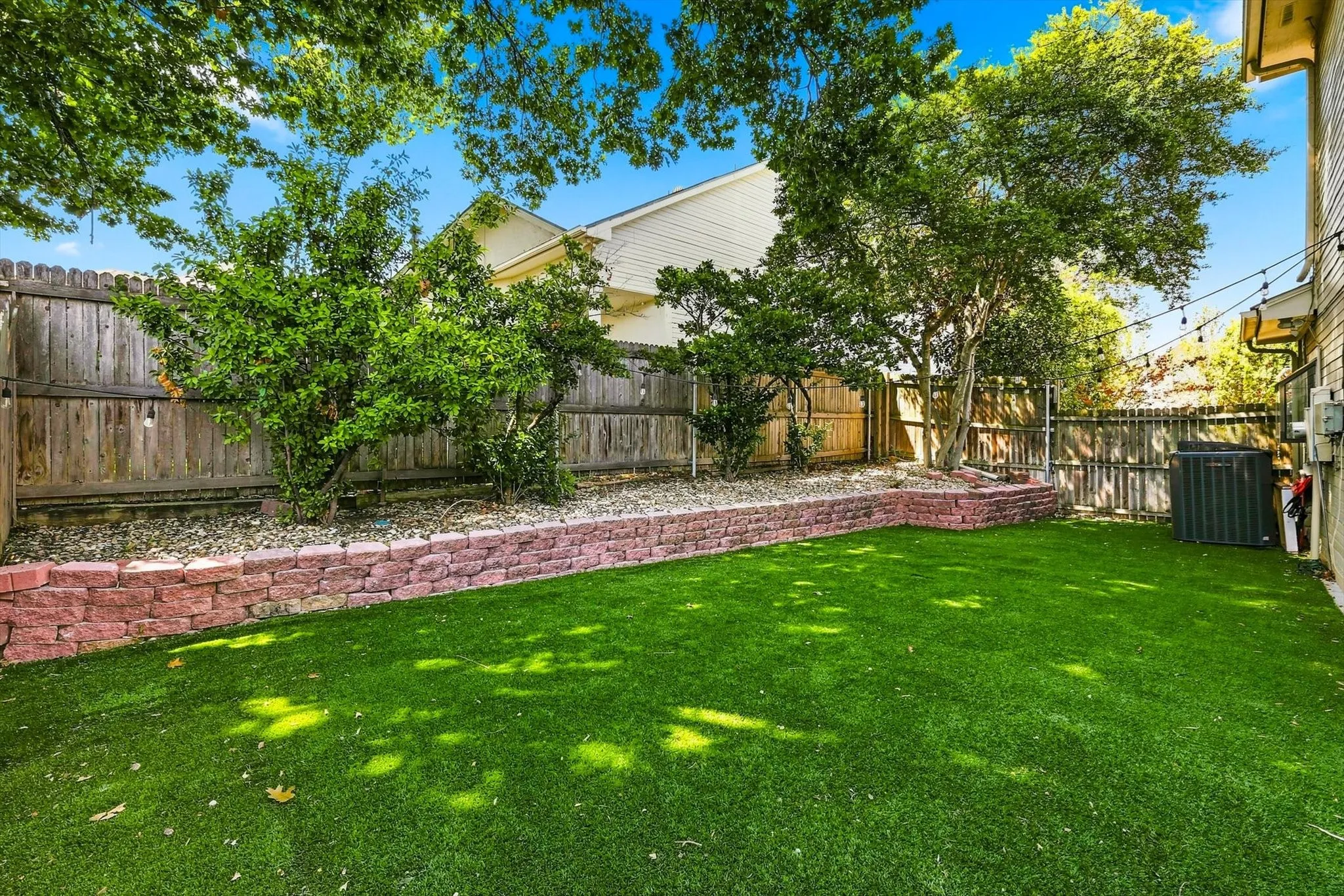 Expansive artificial turf backyard featuring a decorative brick retaining wall, wood fencing, and mature trees