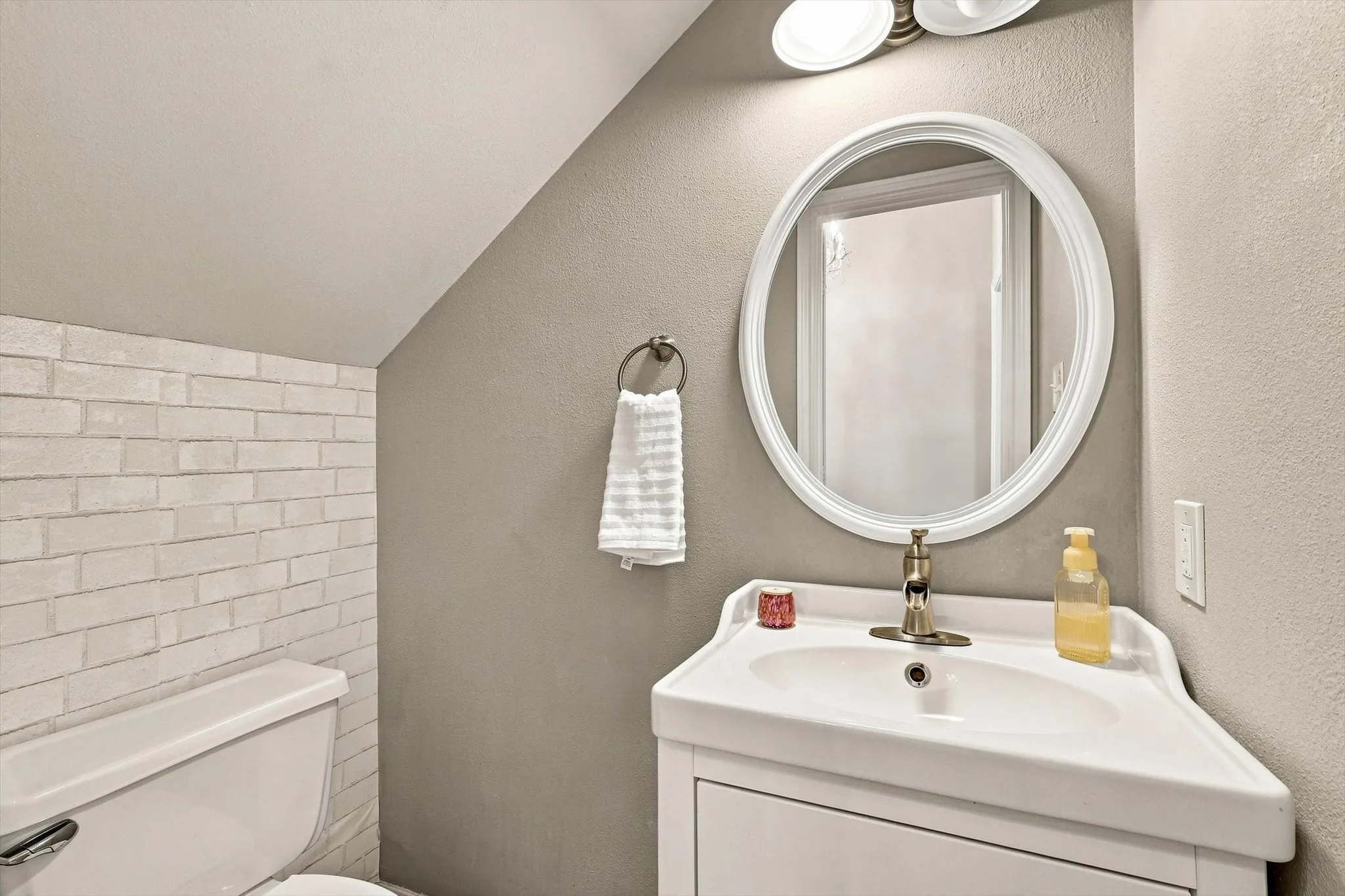 White brick-patterned wall, white vanity with integrated sink, oval framed mirror, and two-light vanity fixture