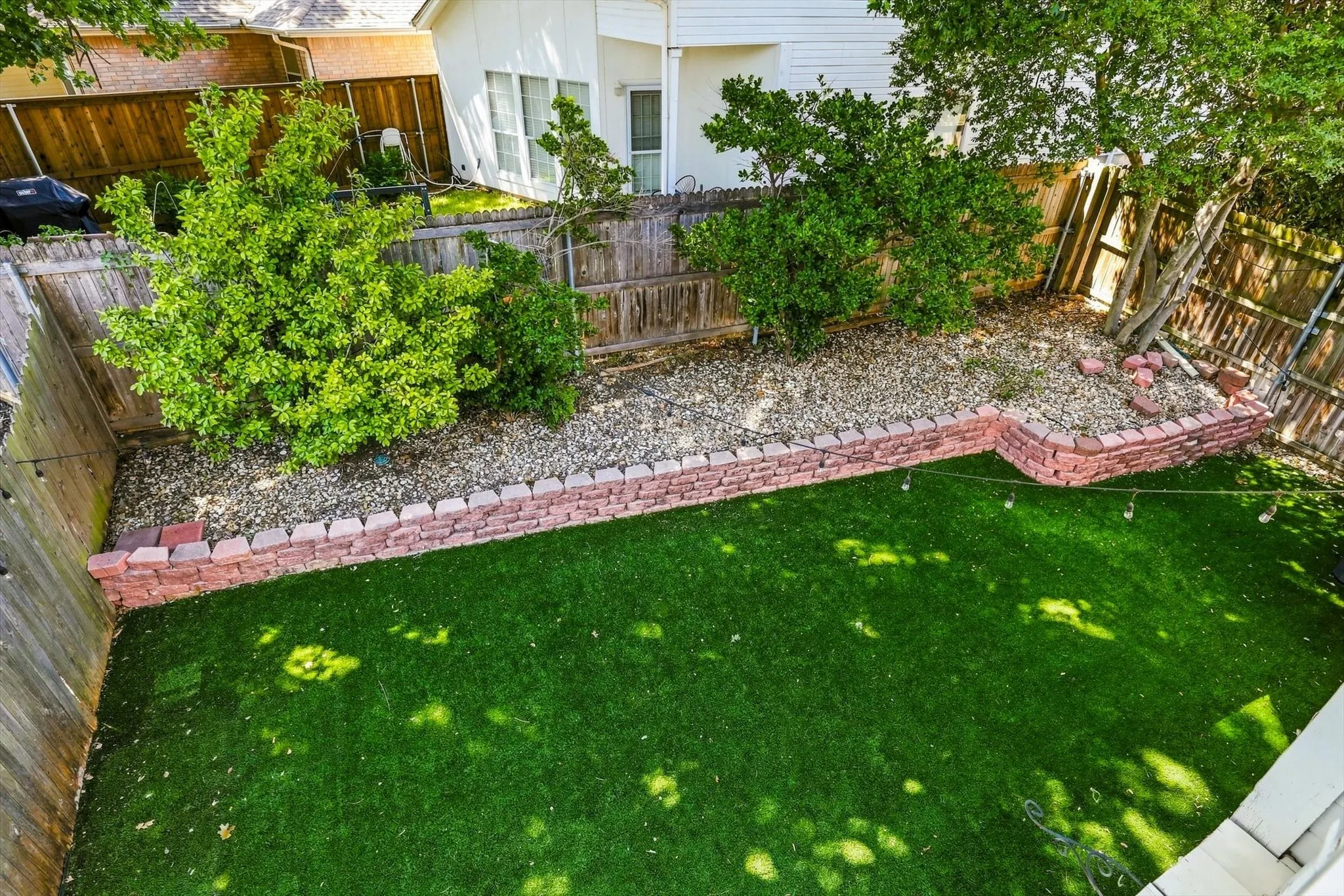 Fenced backyard featuring artificial turf, a decorative brick retaining wall, and mature landscaping with gravel beds