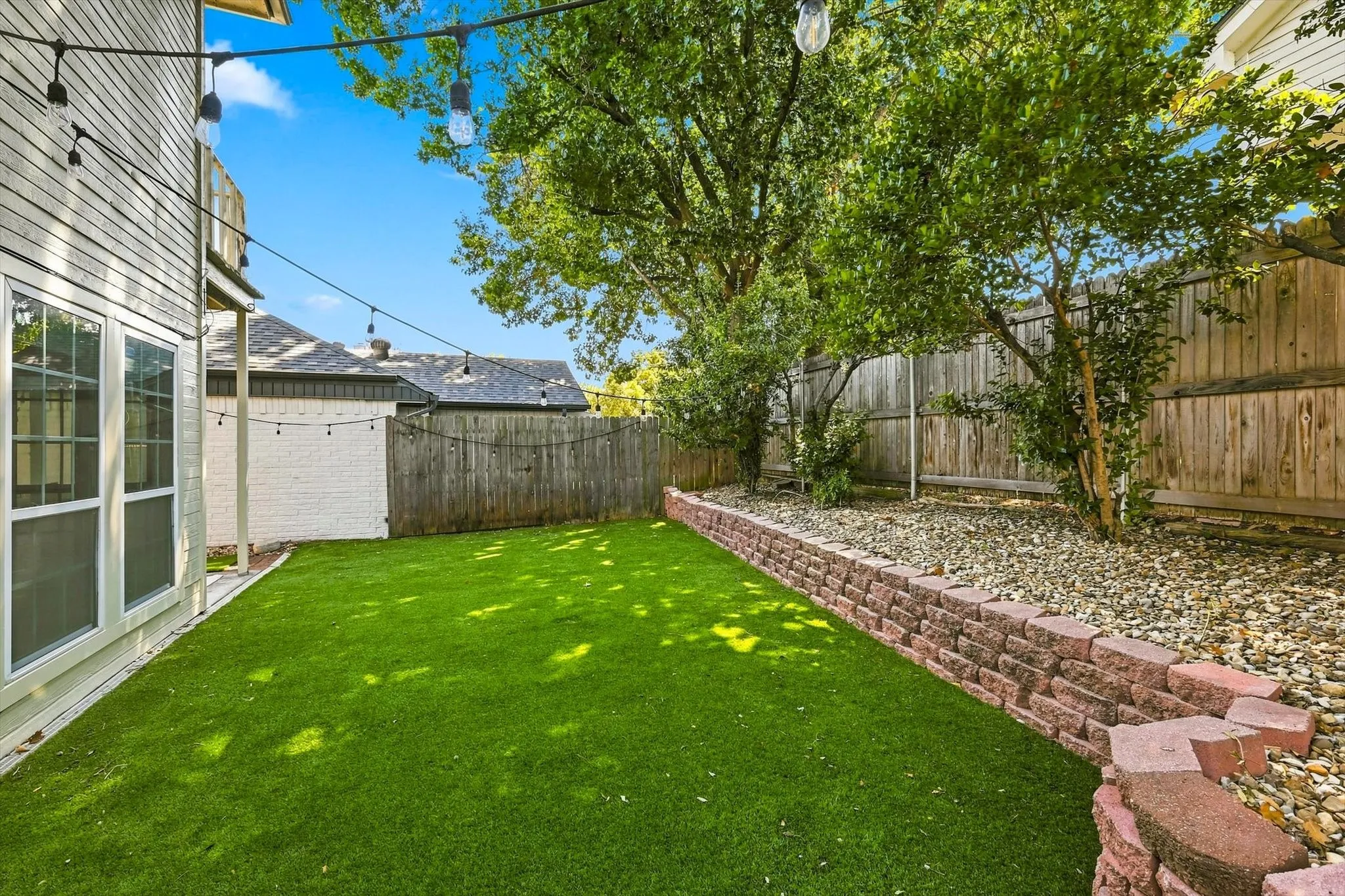 Expansive backyard featuring artificial turf, a decorative brick retaining wall, mature trees, and a privacy fence