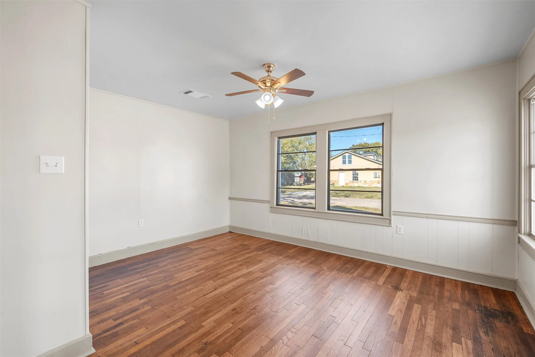 Spacious room featuring wood-finish flooring, white walls with a chair rail and beadboard accent, and a ceiling fan with integrated lighting