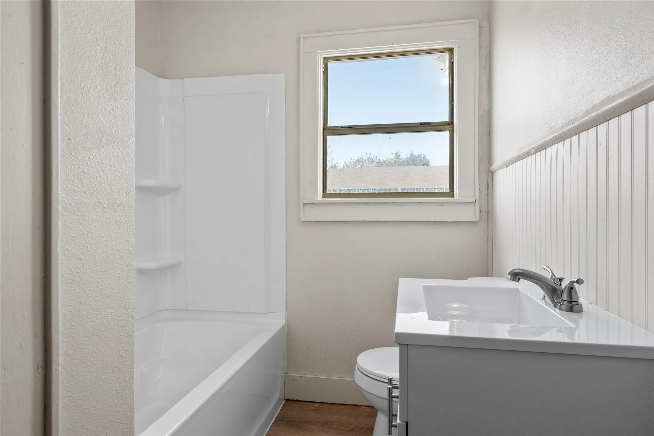 Bathroom featuring a white integrated tub/shower unit, white vanity with an integrated sink, polished chrome faucet, beadboard wainscoting, and wood-finish flooring