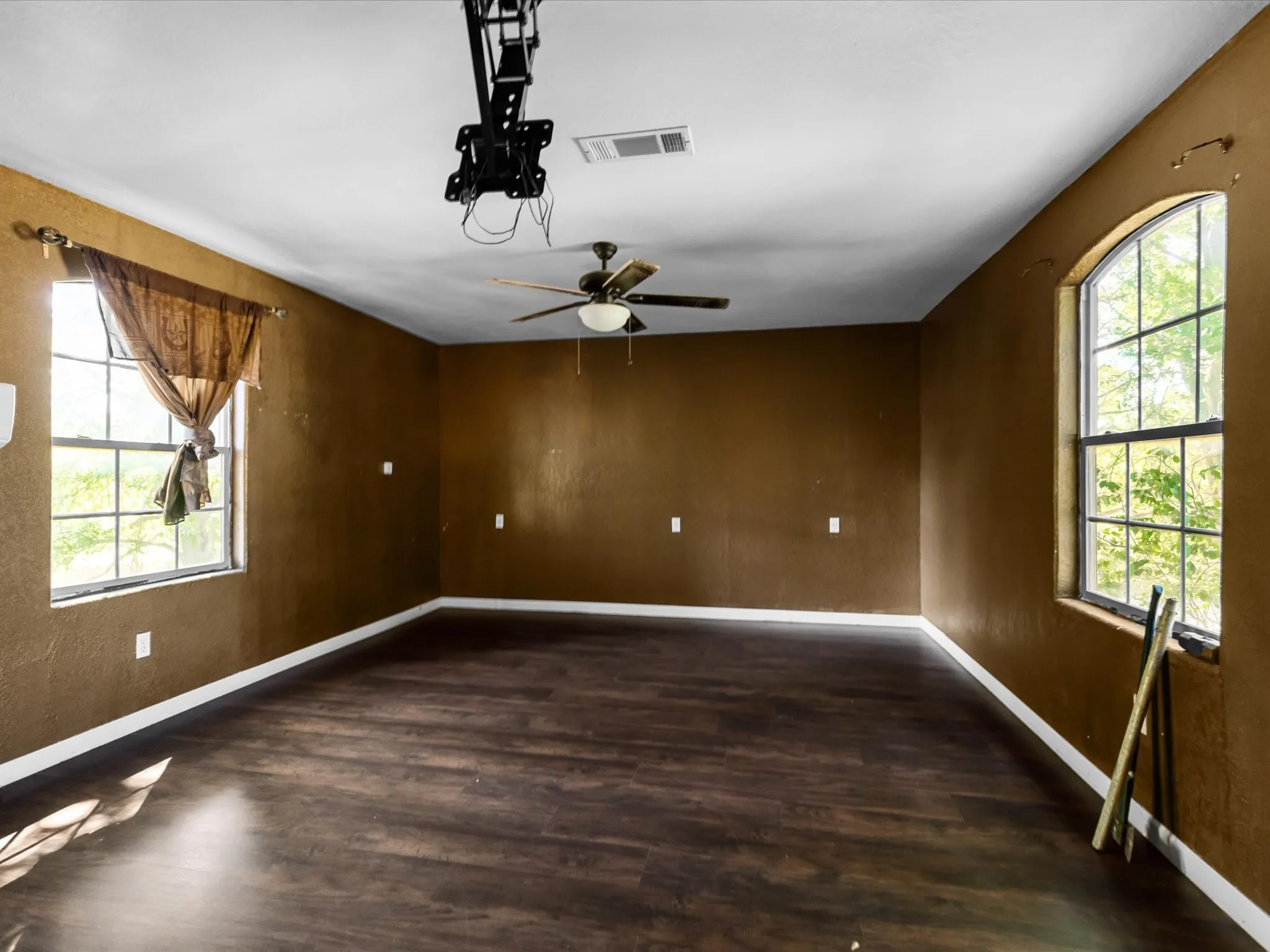 Spacious room featuring dark wood-finish flooring, a ceiling fan, and two large windows including one arched window