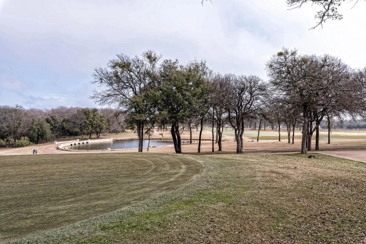 Expansive green space featuring a pond with an arched stone bridge, mature trees, and landscaping