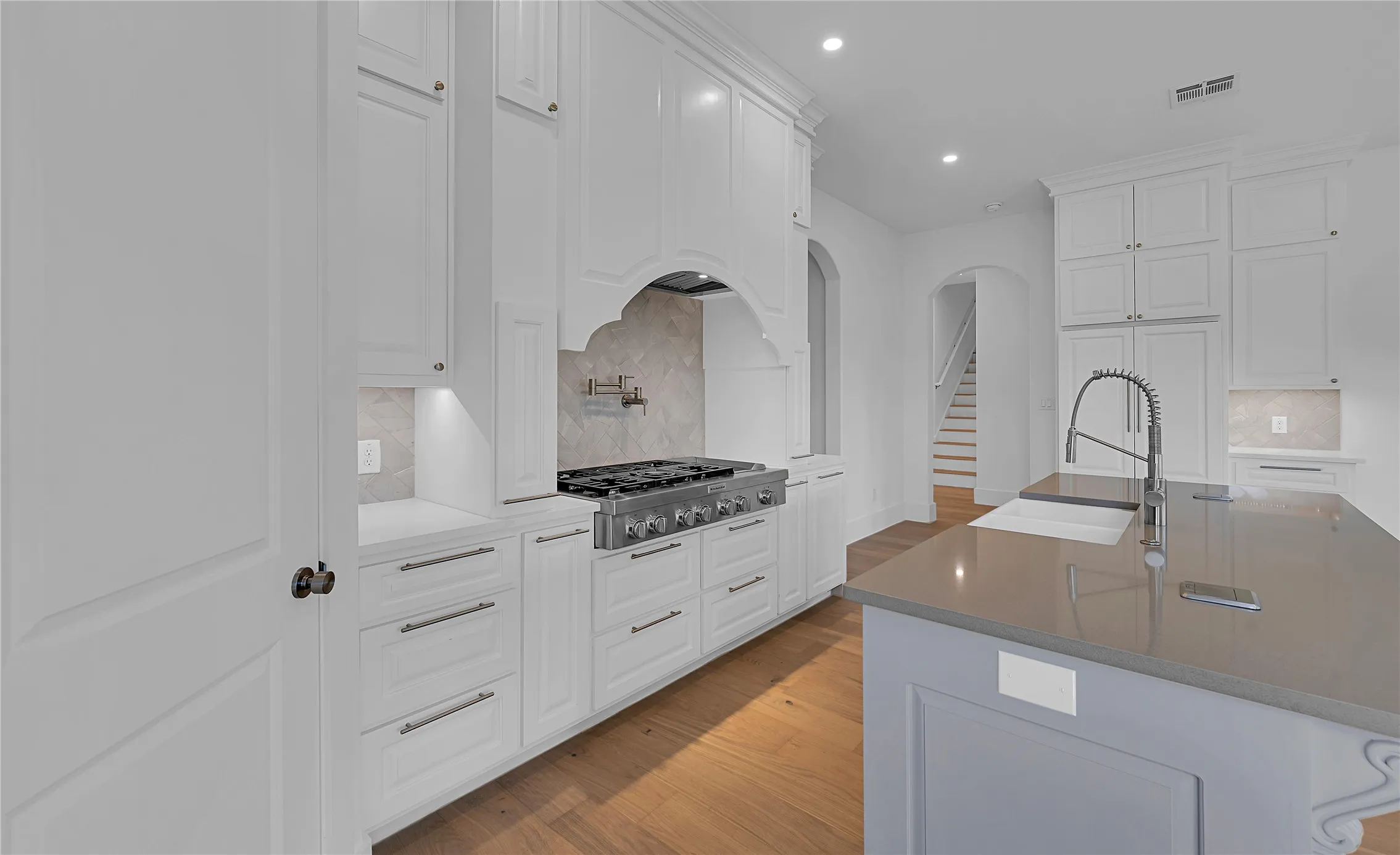 Gourmet kitchen featuring a built-in range, white shaker-style cabinetry, wood-finish flooring, a large island with an undermount sink, and recessed lighting
