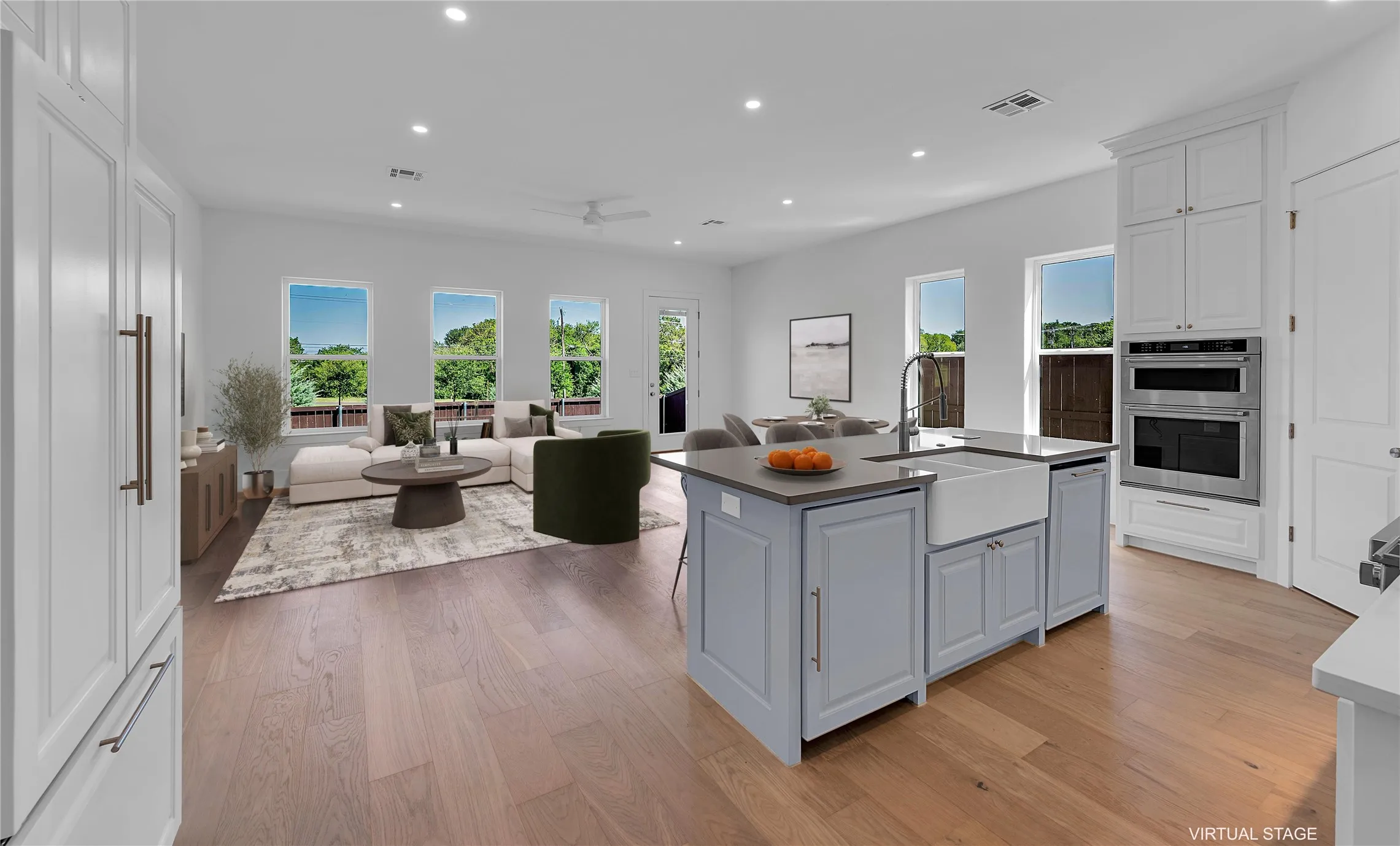 Open-concept living space with wood-finish flooring, a kitchen island with a farmhouse sink, stainless steel built-in double ovens, and multiple windows