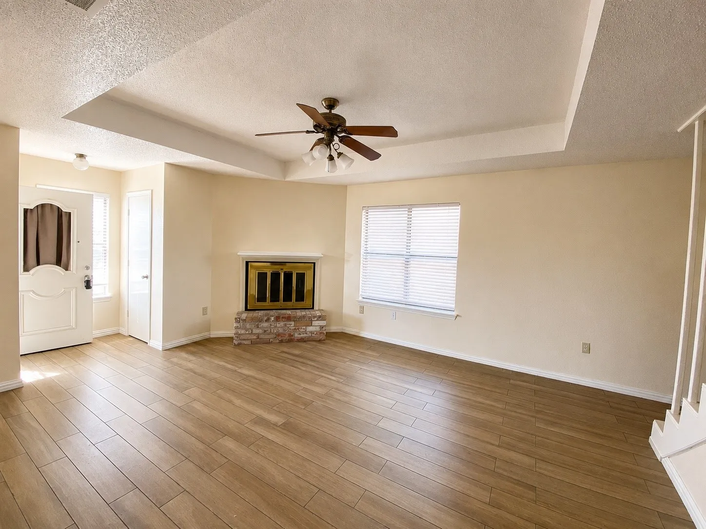 Spacious interior featuring wood-finish flooring, a tray ceiling with a ceiling fan, and a fireplace with a brick base