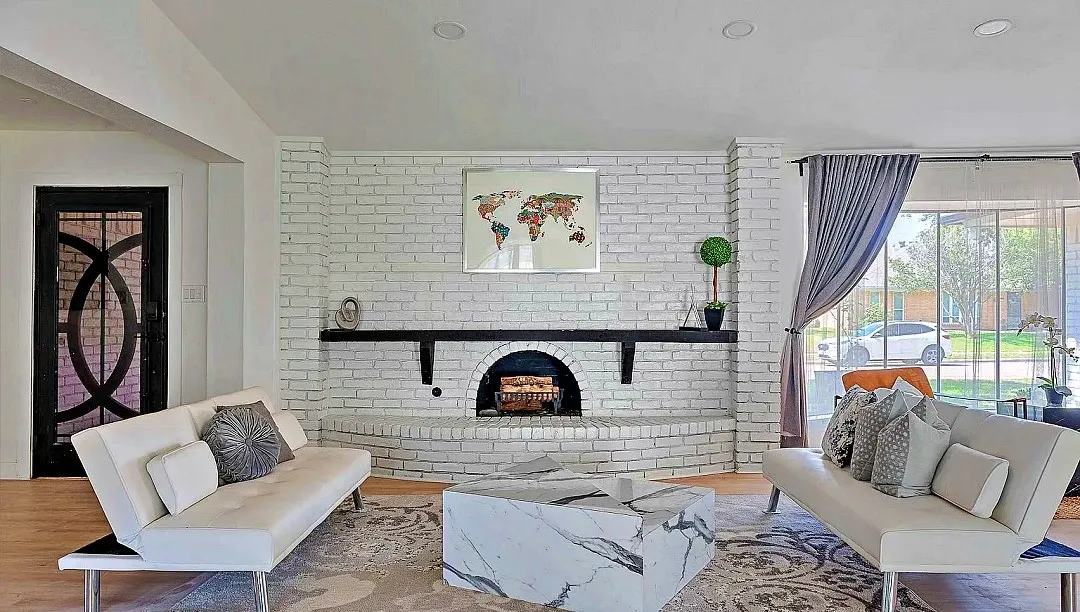 Living room featuring a prominent white brick fireplace with a black mantel, wood-finish flooring, and a decorative black entry door with intricate glasswork
