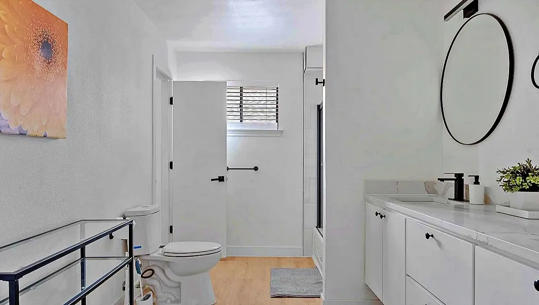 Contemporary bathroom featuring a white vanity with a light-colored countertop, round black-framed mirror, and wood-finish flooring
