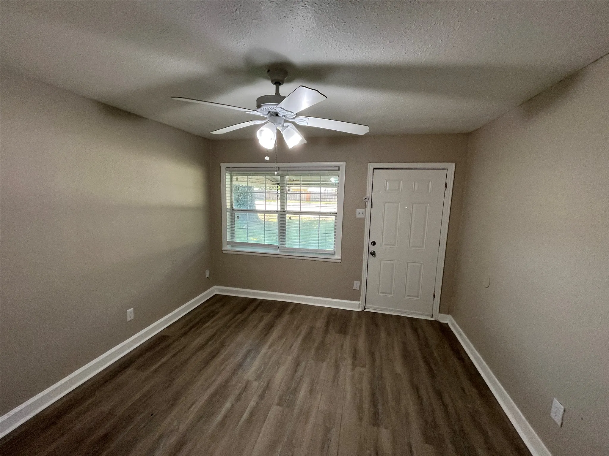 Spacious room featuring wood-finish flooring, a ceiling fan with integrated lighting, a window with horizontal blinds, a paneled entry door, and painted walls with white trim