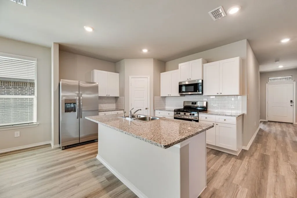 Kitchen featuring a central island with a granite countertop and integrated double basin sink, stainless steel appliances, white shaker cabinetry, subway tile backsplash, and wood-finish flooring
