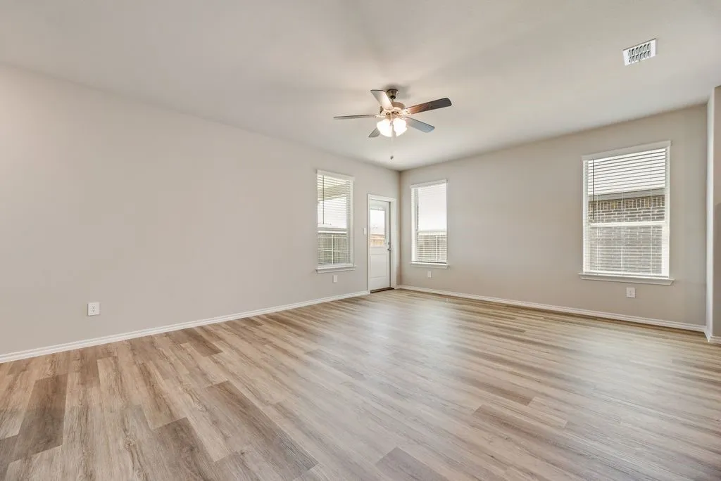 Spacious room featuring wood-finish flooring, light neutral wall tones, multiple windows with blinds, a ceiling fan with integrated lighting, and a white door with a window