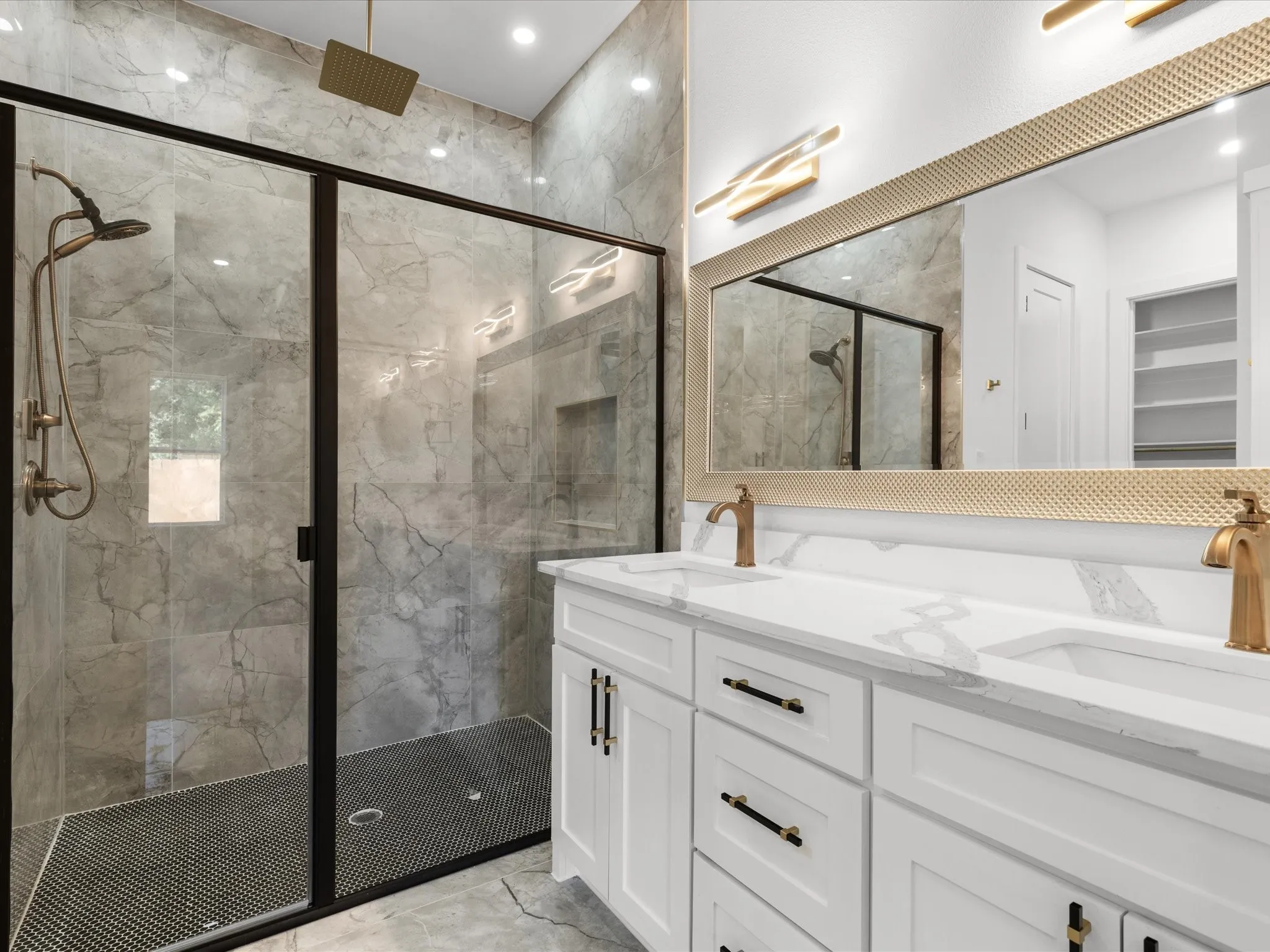 Spacious bathroom featuring a glass-enclosed shower with large format tile surround and rainfall showerhead, dual vanity with white cabinetry, quartz-finish countertops, and brass-finish fixtures
