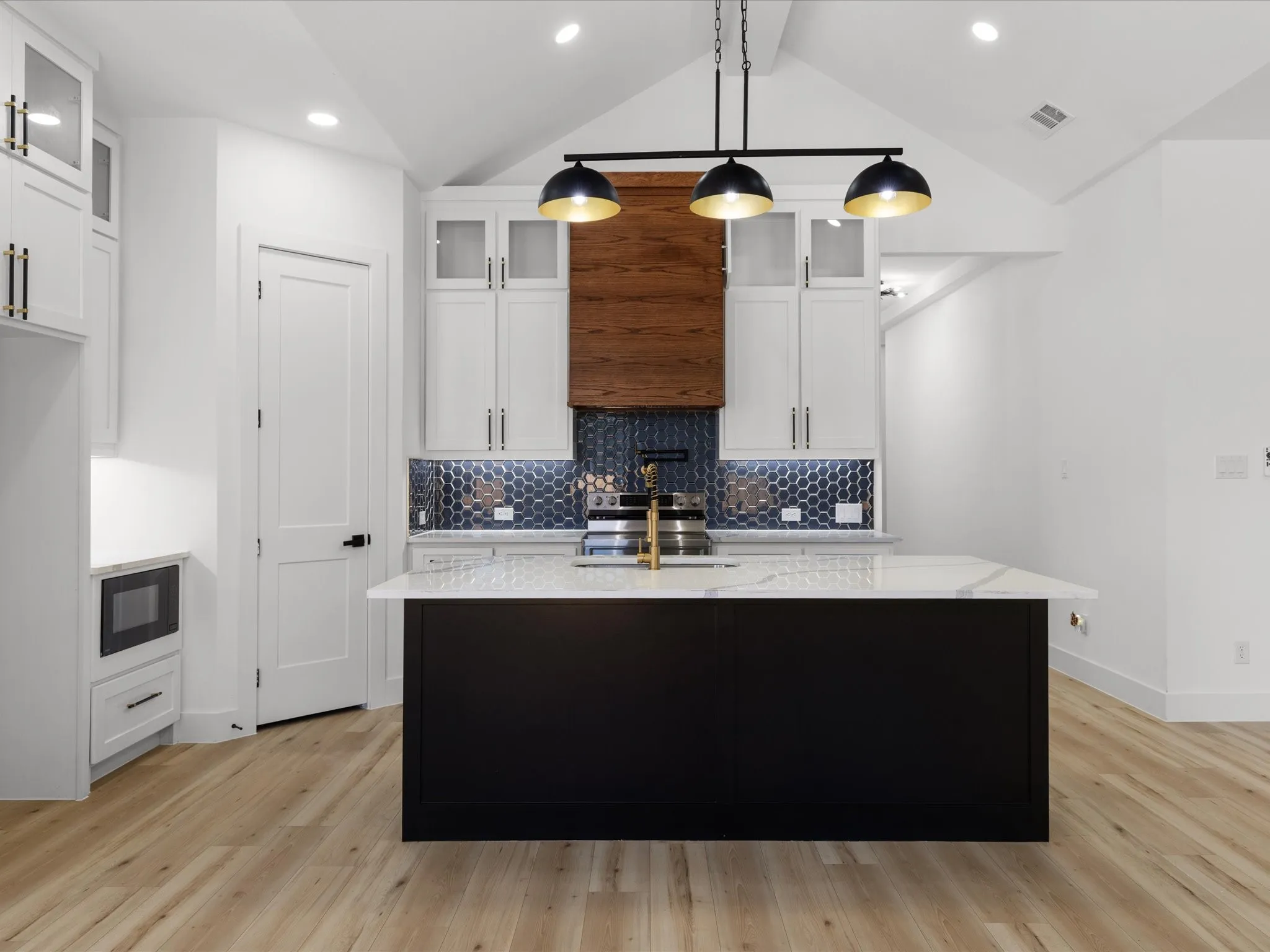 Modern kitchen featuring a large central island with a light-toned countertop, white cabinetry with gold-toned hardware, and a dark hexagonal tile backsplash