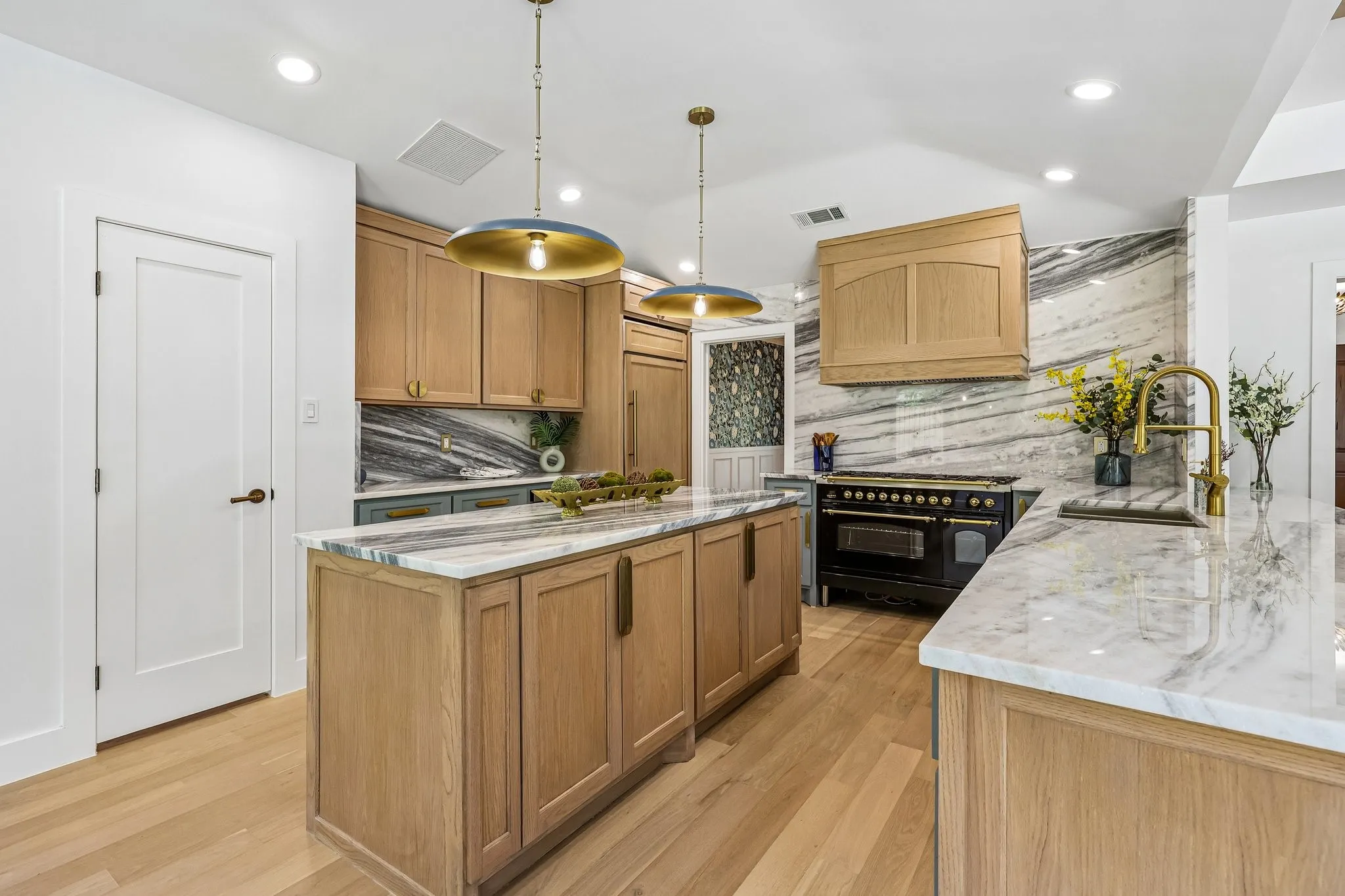 Spacious kitchen featuring light wood-finish cabinetry, a large center island with a stone countertop, a built-in range with a wood-finish hood, and statement pendant lighting