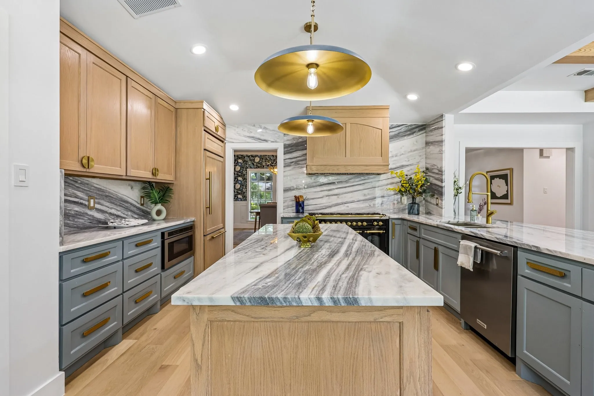 Kitchen featuring a large central island with a stone countertop, light wood cabinetry, gray base cabinetry with gold-toned hardware, full-height stone backsplash, and recessed lighting