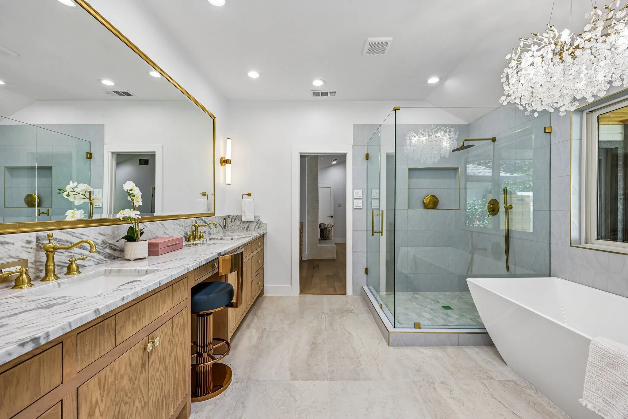 Spacious bathroom featuring a dual vanity with a patterned stone countertop and brass fixtures, a frameless glass shower enclosure with a rainfall showerhead, a freestanding soaking tub, and a decorative chandelier