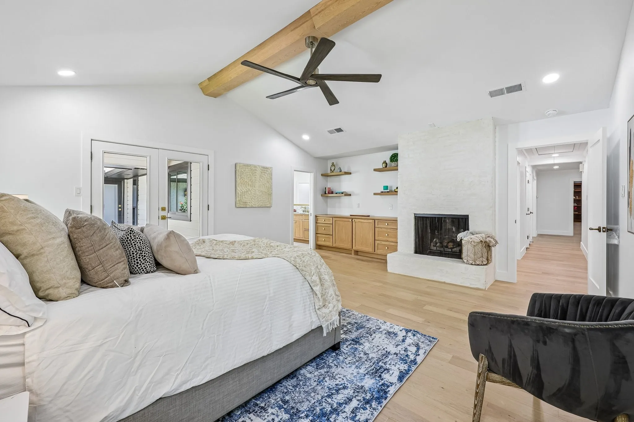 Vaulted ceiling featuring a wood beam and ceiling fan, white painted brick fireplace, built-in cabinetry with wood-finish shelving, light wood-finish flooring, and double glass-panel doors