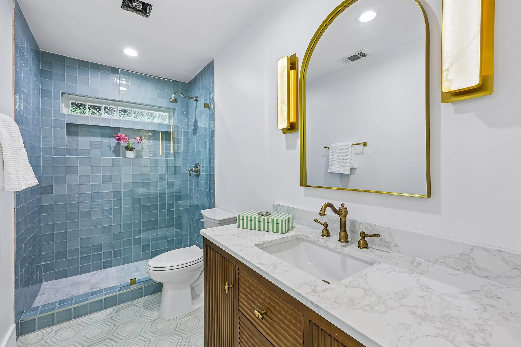 Contemporary bathroom featuring a wood-finish vanity with a white countertop and undermount sink, brass-tone hardware, and an arched mirror with flanking sconces