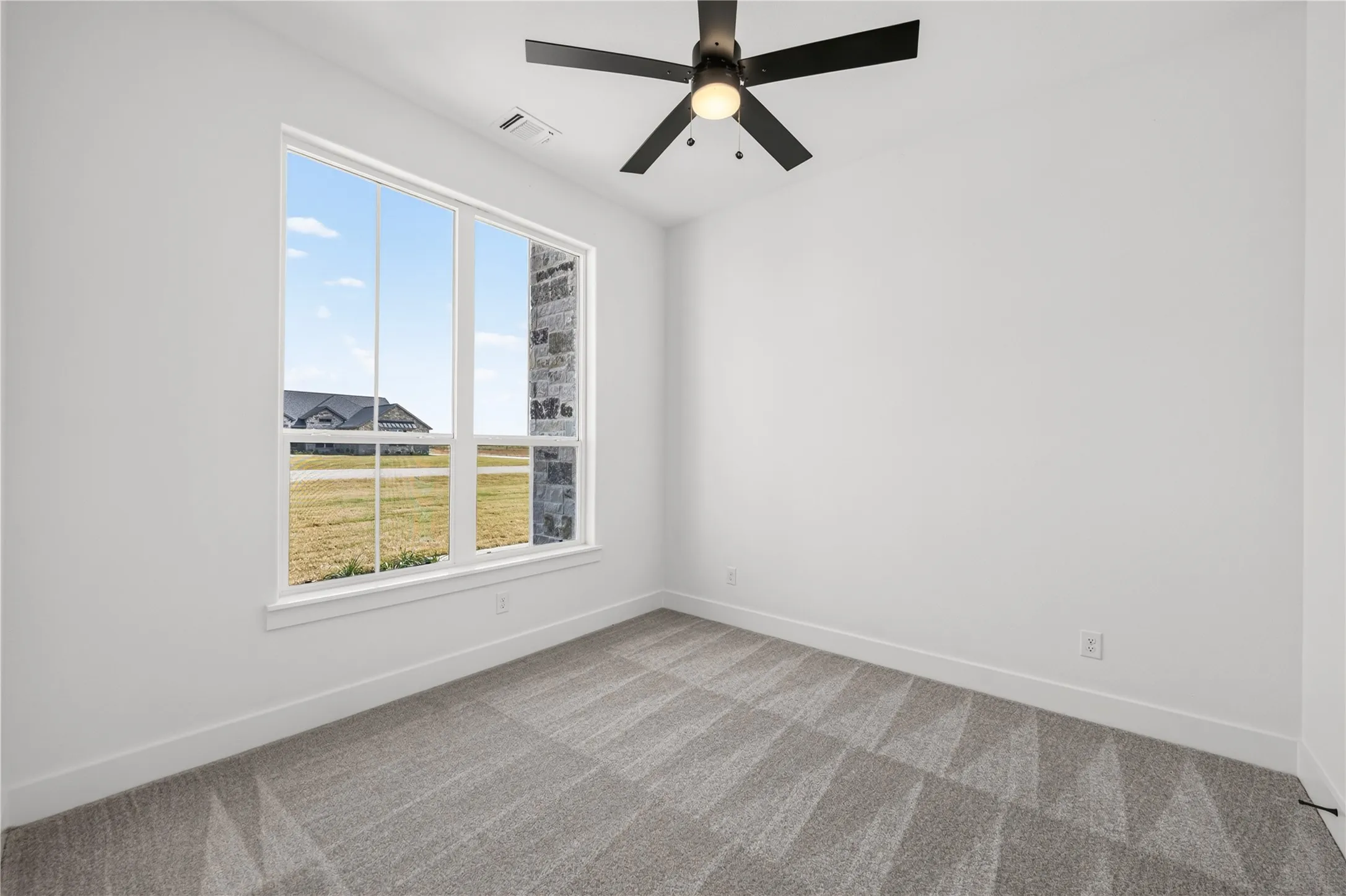 Room featuring a large multi-pane window, patterned grey carpet, white baseboards, and a ceiling fan with light fixture