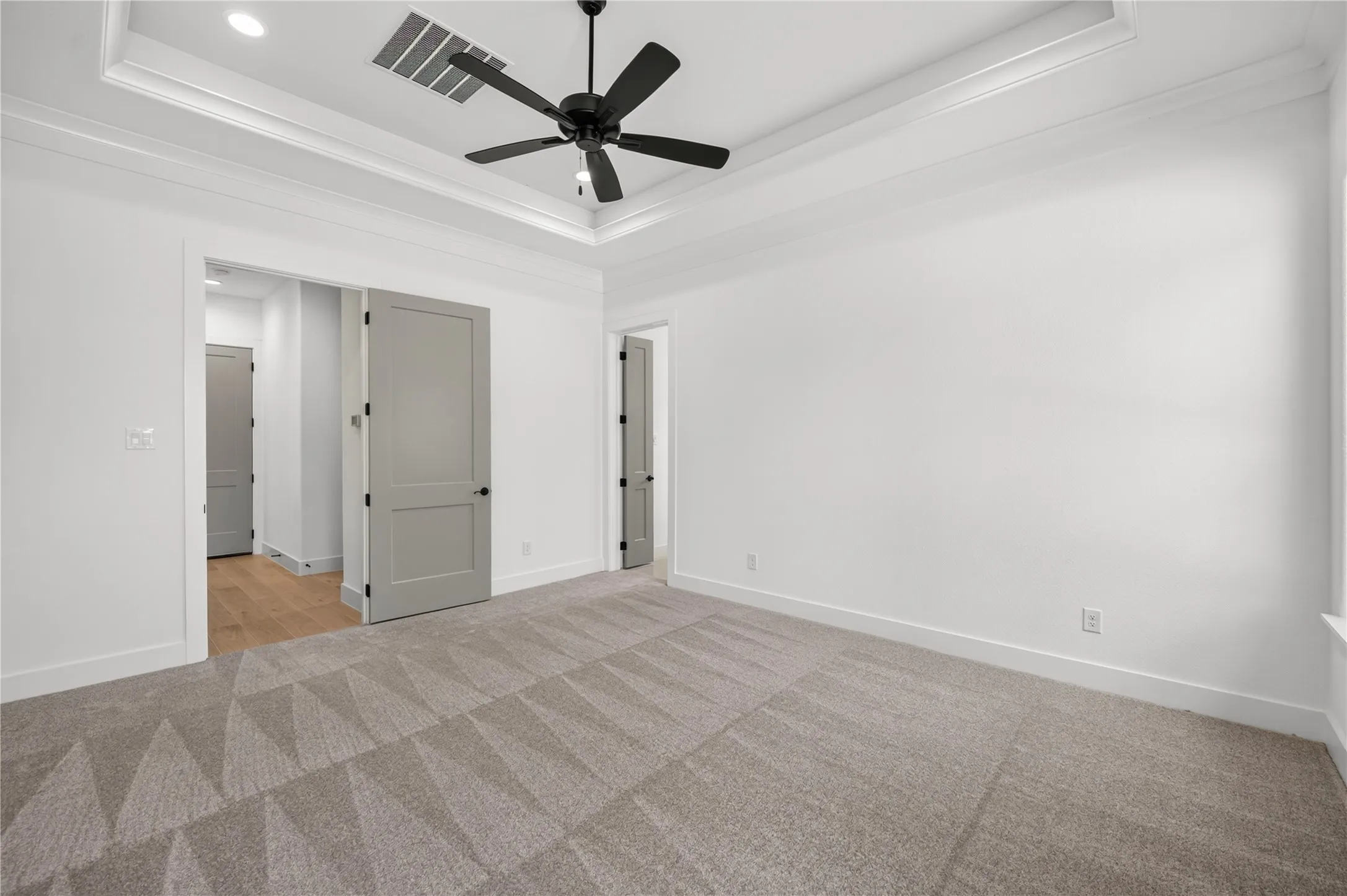 Spacious room featuring a tray ceiling with a black ceiling fan, light gray patterned carpeting, and white walls
