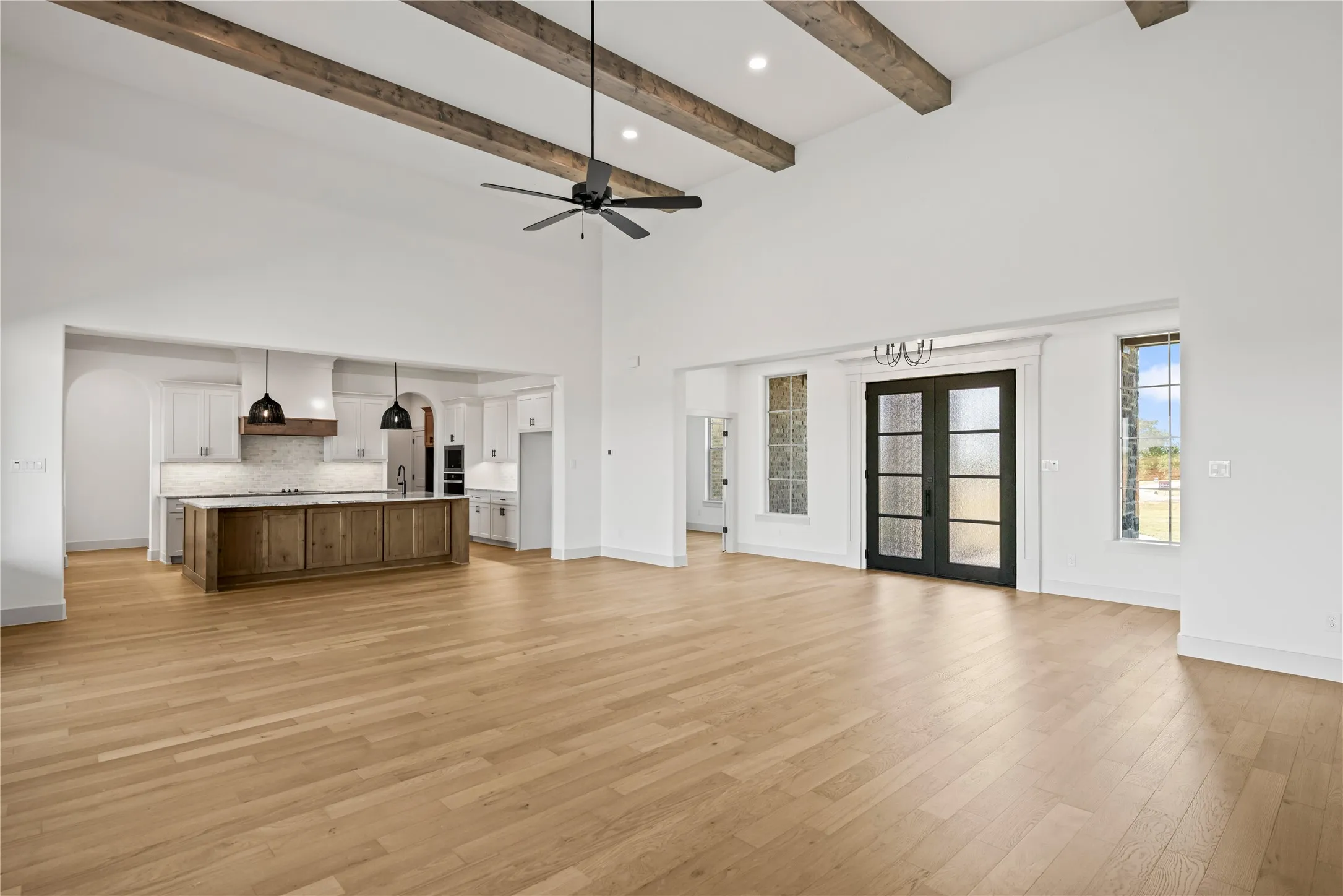 Spacious open-concept living area featuring vaulted ceilings with exposed wood beams, light wood-finish flooring, and a contemporary ceiling fan