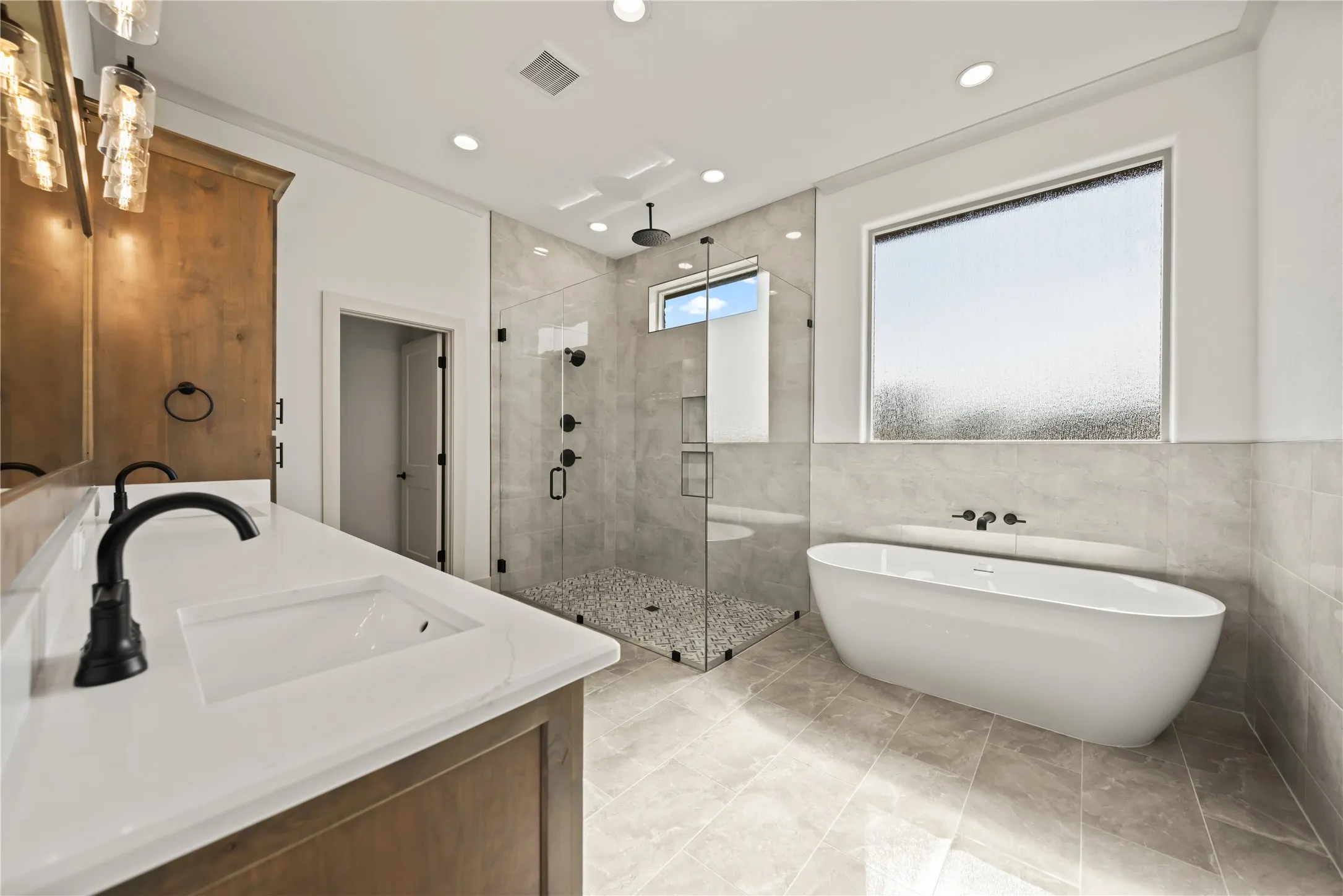 Spacious bathroom featuring a freestanding soaking tub, a frameless glass shower enclosure with pebble flooring, and a dual vanity with white countertops and matte black fixtures