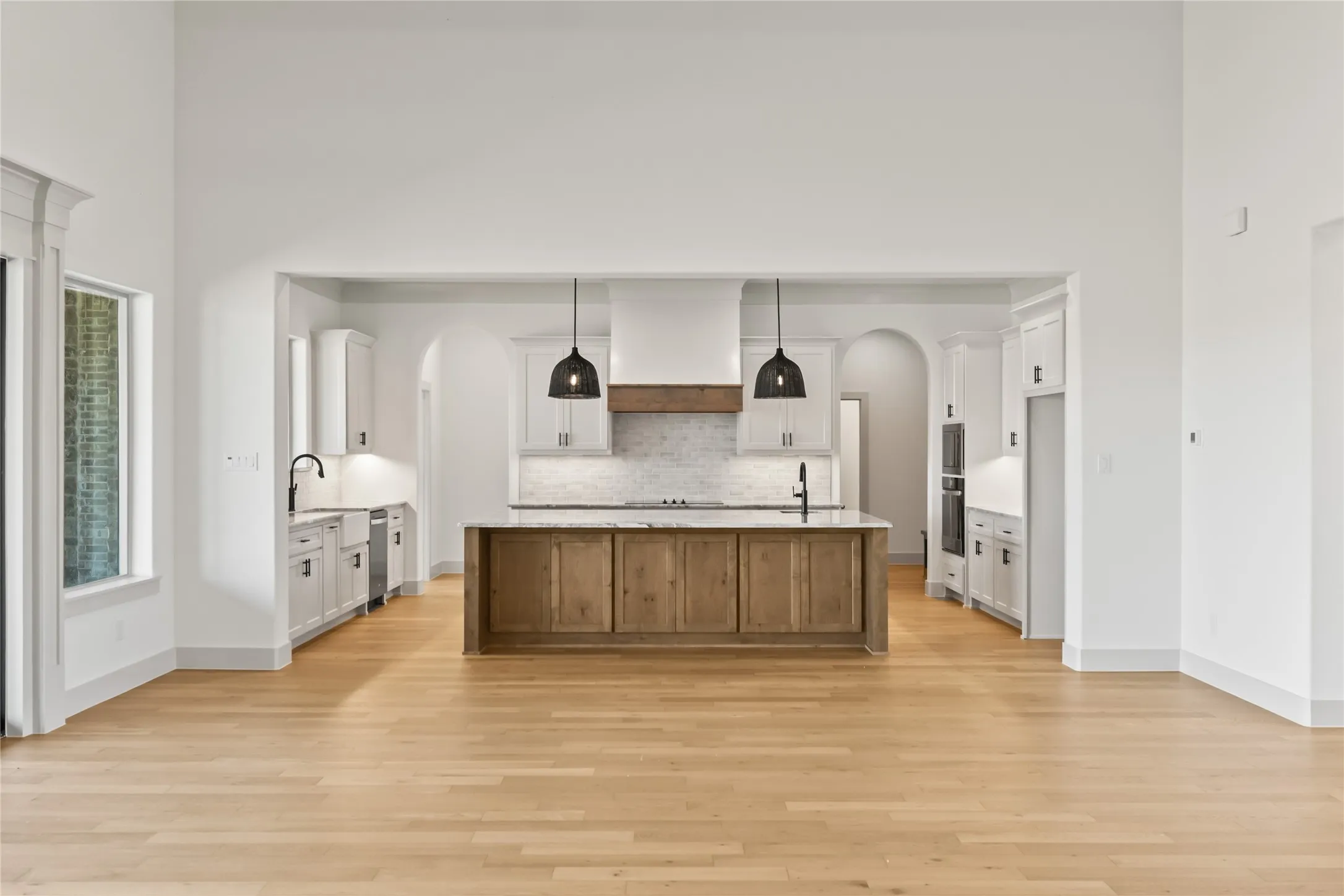 Open-concept kitchen featuring light wood-finish flooring, a wood-paneled island with a light-toned countertop, white cabinetry with dark hardware, a farmhouse sink, and a built-in range hood with a wood accent
