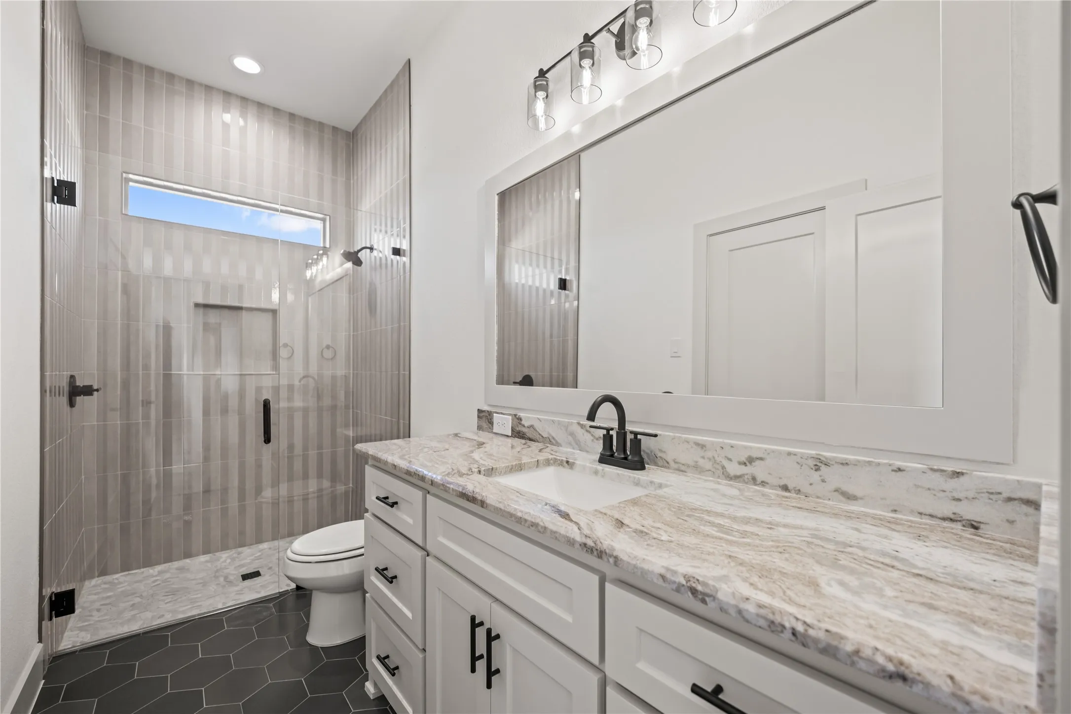 Bathroom featuring a frameless glass shower enclosure, a transom window, floor-to-ceiling vertical tile, a built-in shower niche, and a vanity with a stone countertop