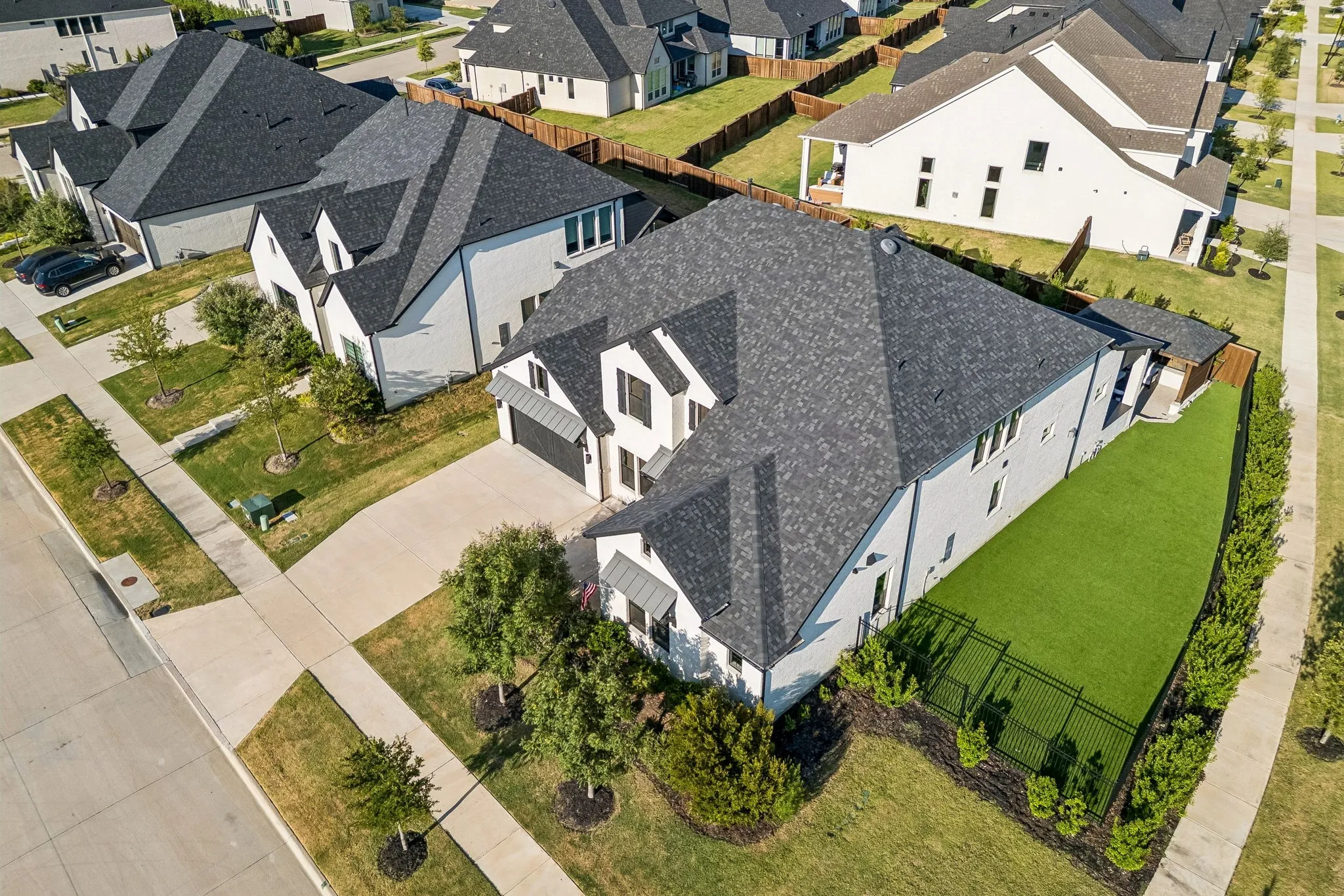 Contemporary white exterior with a dark shingle roof, featuring a fenced side yard with artificial turf, an attached garage, and established landscaping