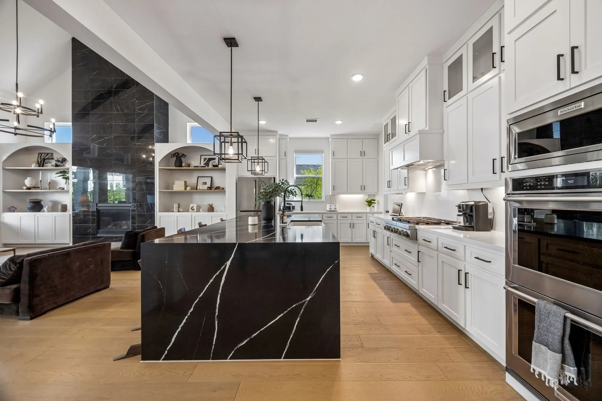 Open-concept kitchen and living area featuring a large black waterfall island, white cabinetry with black hardware, stainless steel appliances, and wood-finish flooring