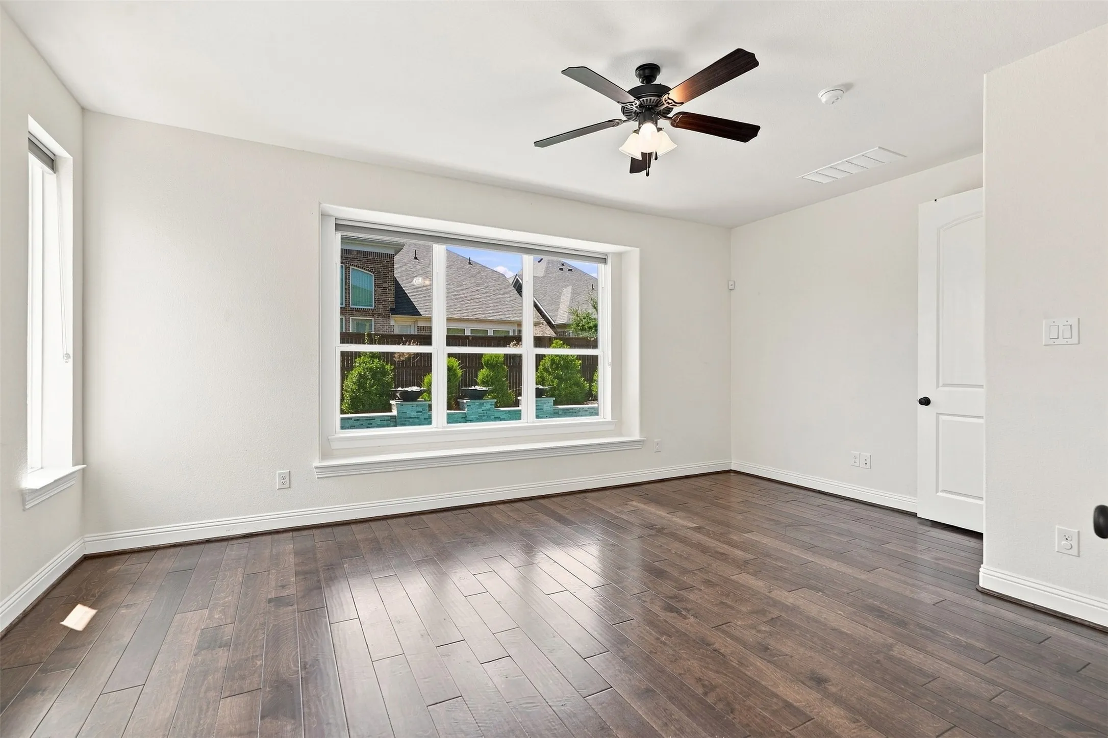 Spacious room featuring rich wood-finish flooring, a multi-pane window, and a ceiling fan with integrated lighting