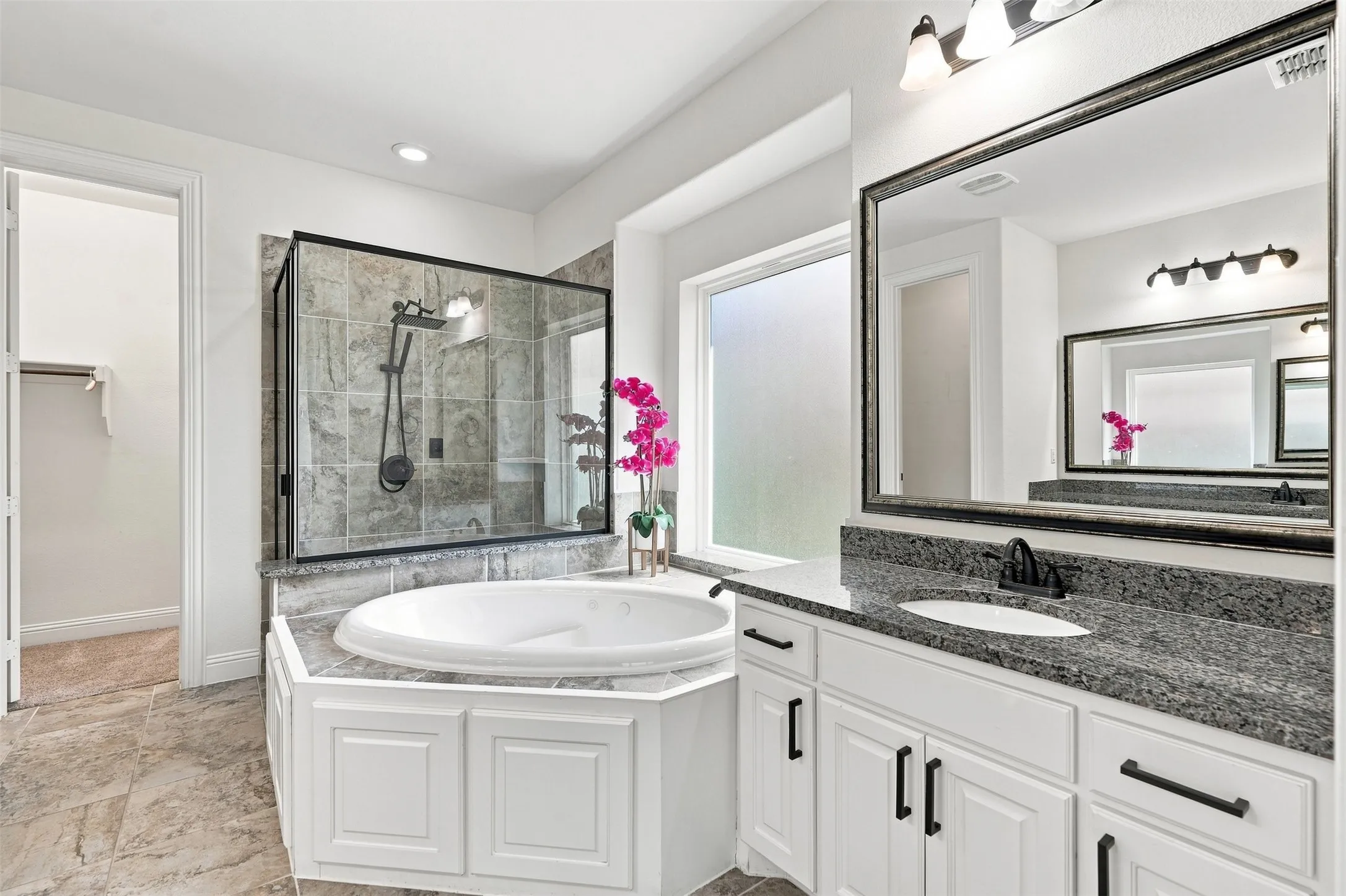Bathroom featuring a separate walk-in shower with tile surround, a built-in garden tub, a double vanity with granite countertops and white cabinetry, and large format tile flooring