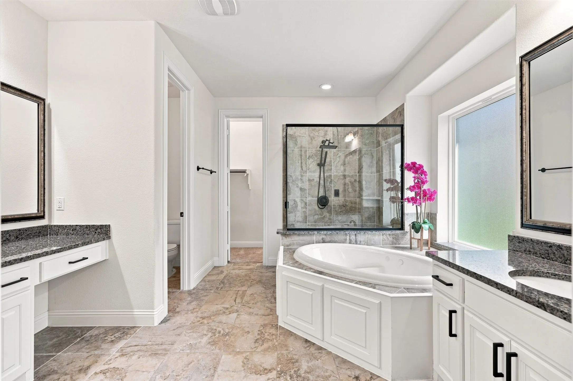 Spacious bathroom featuring a built-in soaking tub, a glass-enclosed shower, dual vanities with dark countertops, and tile flooring