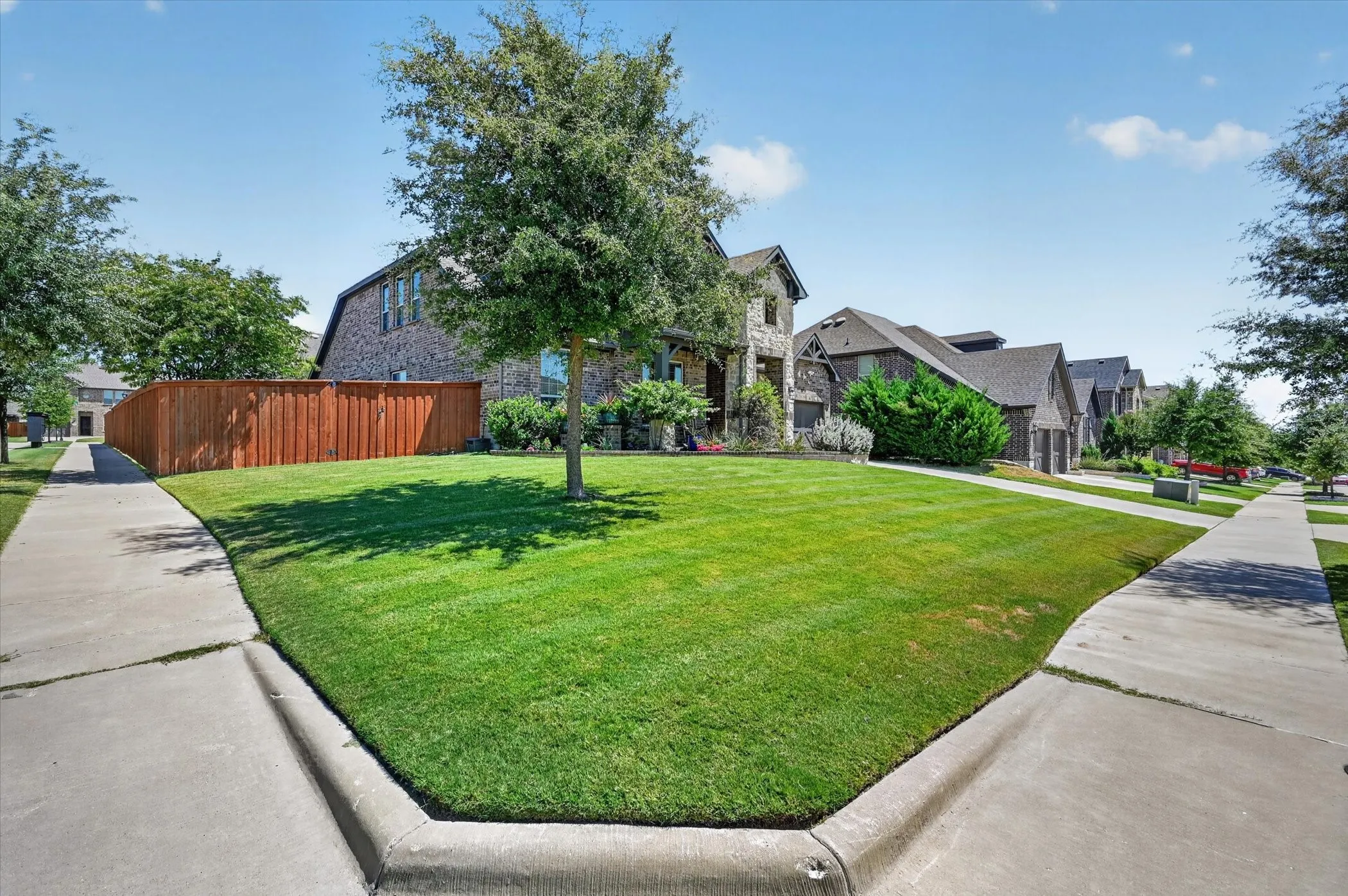 Expansive corner lot featuring a manicured lawn, established trees, concrete sidewalks, and a wood privacy fence