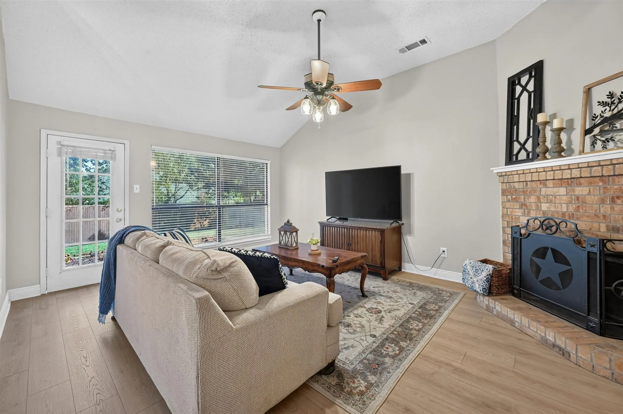 Living area featuring a vaulted ceiling with a ceiling fan, wood-finish flooring, a brick fireplace with a white mantel, and a glass-paned exterior door