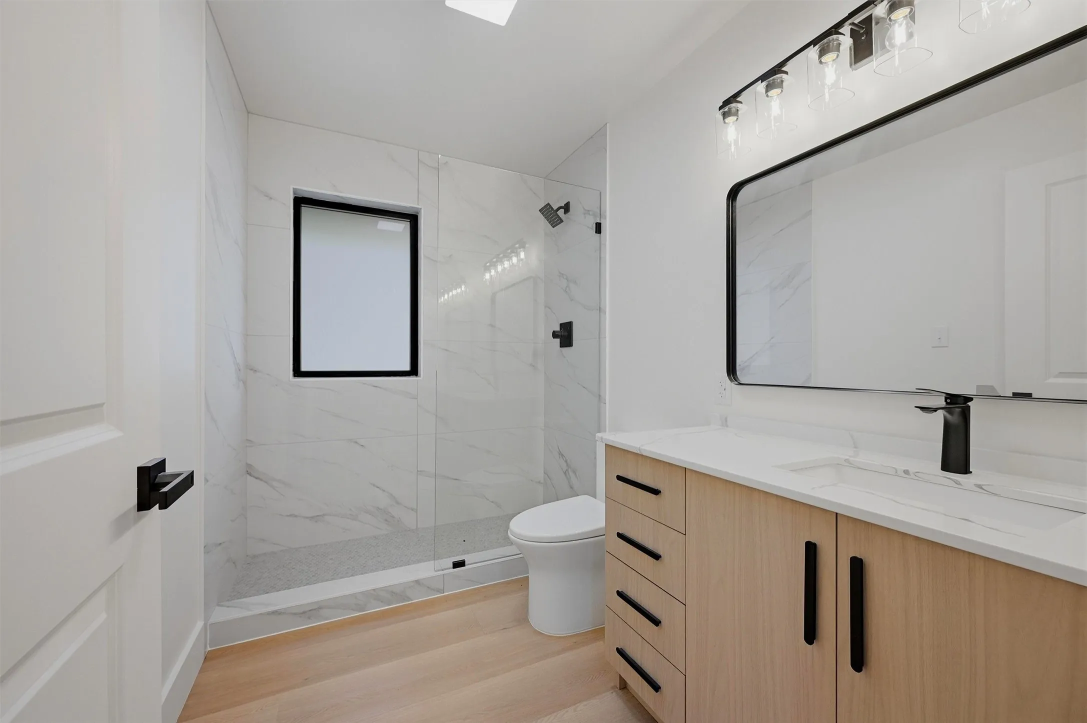 Modern bathroom featuring a light wood-finish vanity with matte black hardware, a rectangular vanity mirror, and a contemporary light fixture