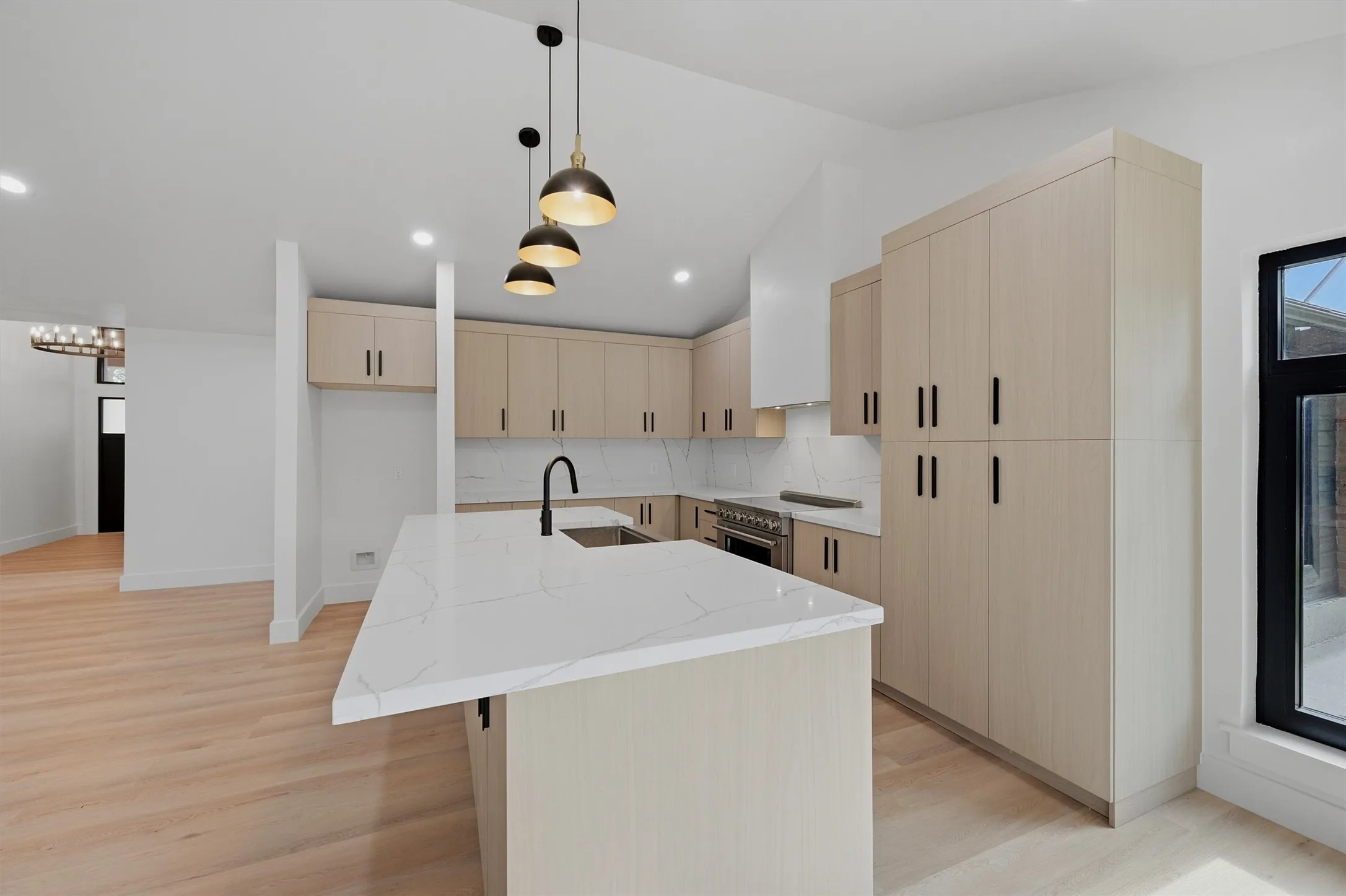 Modern kitchen featuring a large island with a waterfall countertop, light wood-finish cabinetry, stainless steel appliances, and sleek black hardware