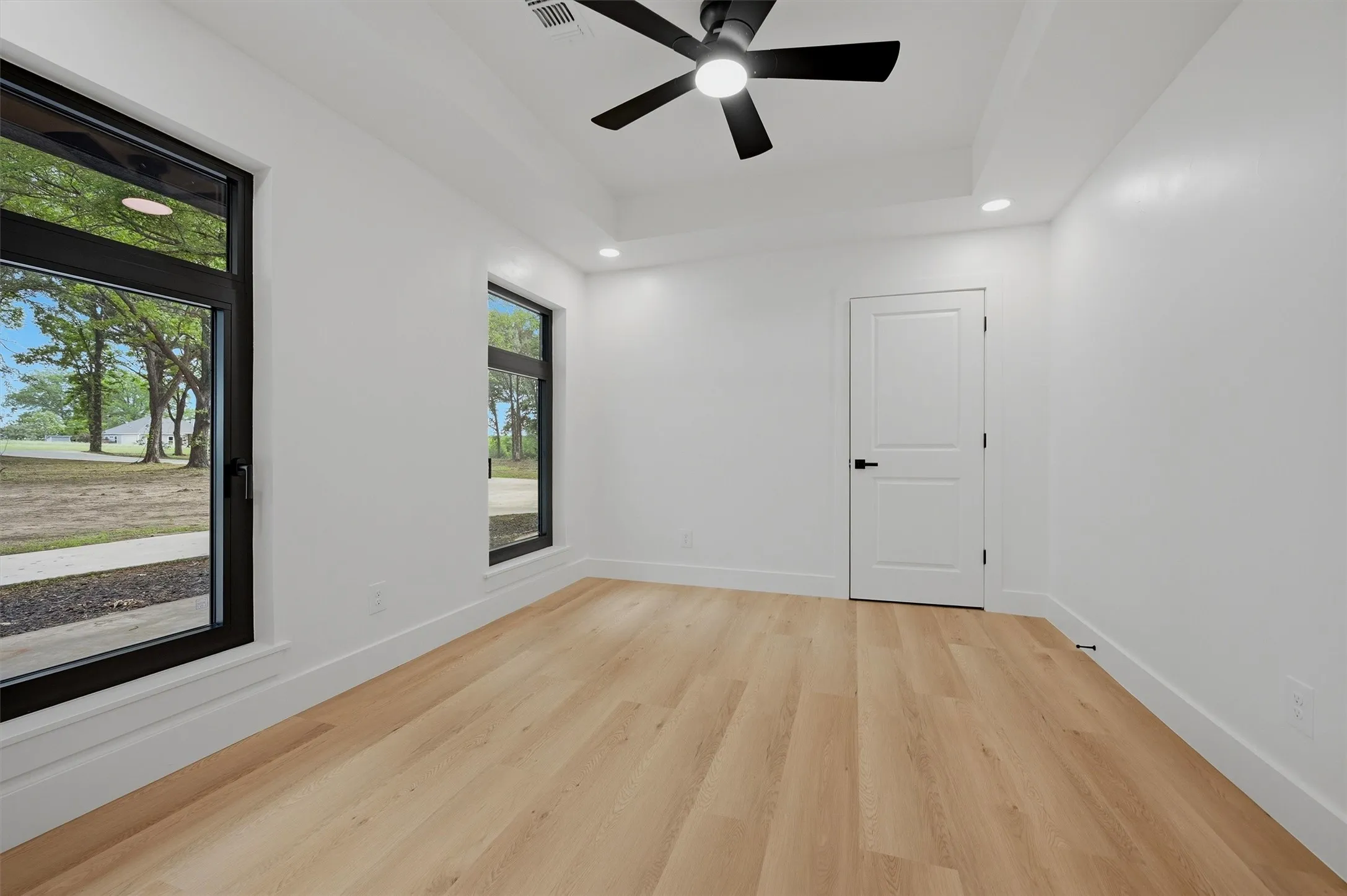 Spacious room featuring light wood-finish flooring, a contemporary ceiling fan, and two large windows with black framing