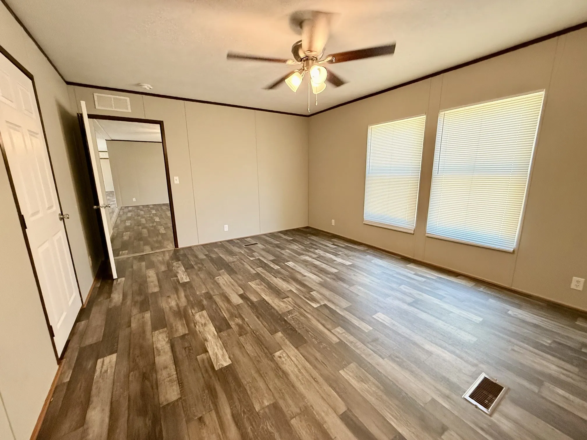 Spacious room featuring wood-finish flooring, two large windows with blinds, a ceiling fan with integrated lighting, and neutral wall tones