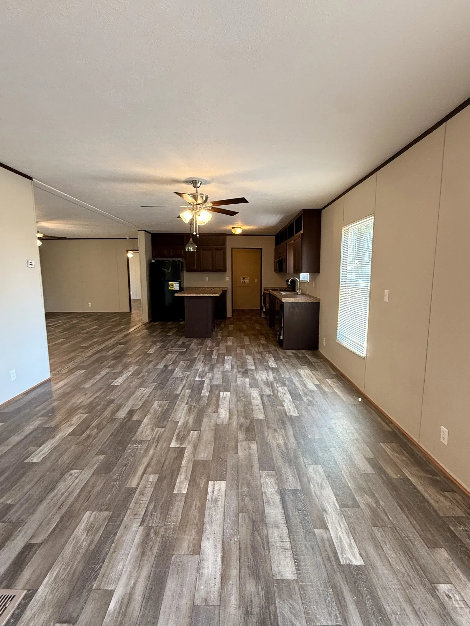 Open-concept interior featuring wood-finish flooring, a ceiling fan with integrated lighting, and a kitchen area with dark cabinetry, a black refrigerator, and a stainless steel sink
