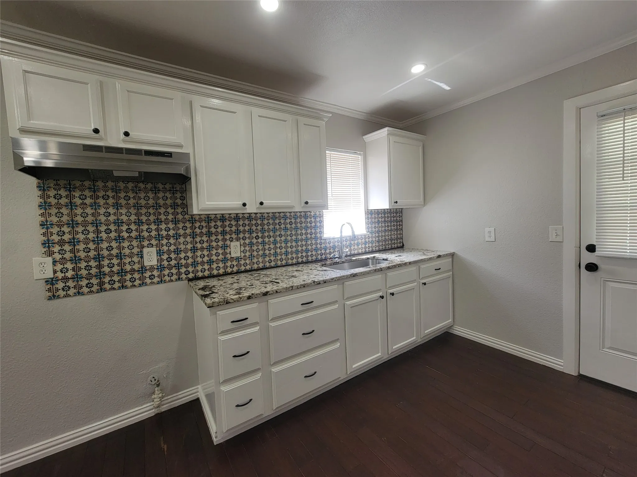 White cabinetry with black hardware, granite-style countertops, mosaic tile backsplash, and dark wood-finish flooring