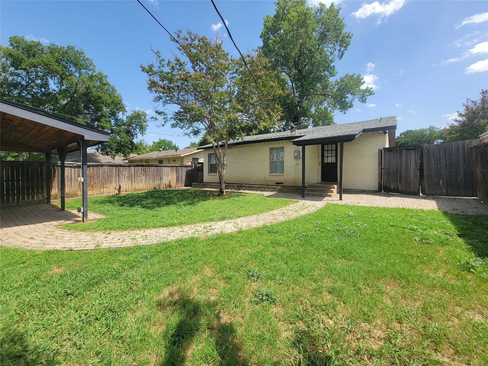 Spacious backyard featuring a covered patio, brick paver pathways, perimeter wood fencing, and mature trees